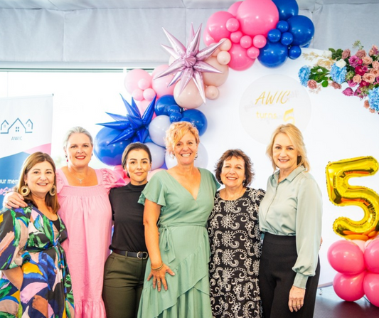 A group of six pose in front of a colorful balloon arrangement, floral decorations, and a sign that reads 'AWIC.' A large gold '5' balloon suggests a 5-year anniversary celebration for AWIC. Prop hire, Prop hiring, Prop hire, event stylists Brisbane, brisbane event styling, brisbane event styling, event stylist Brisbane, Brisbane Wedding, Gold Coast Wedding, Floral Styling, Lush & Lavish Events, Brisbane Tablescape, Gold Coast Tablescapes, Brisbane Candle Hire