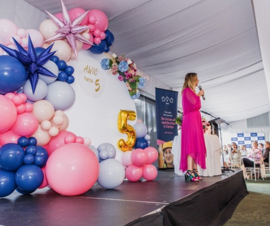 Person in a long pink dress speaking on a stage decorated with pink, blue, white, and purple balloons, large metallic number ‘5’, and the word ‘AMIC’. Audience facing the stage, backs to the camera. Prop hire, Prop hiring, Prop hire, event stylists Brisbane, brisbane event styling, brisbane event styling, event stylist Brisbane, Brisbane Wedding, Gold Coast Wedding, Floral Styling, Lush & Lavish Events, Brisbane Corporate Styling, Gold Coast Corporate Styling, Brisbane Brand Activation