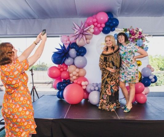 Two people standing in front of a colorful balloon backdrop. One takes a selfie with a smartphone, while the other stands beside them. Pink, blue, and purple balloons with floral decorations suggest a celebratory event. Prop hire, Prop hiring, Prop hire, event stylists Brisbane, brisbane event styling, brisbane event styling, event stylist Brisbane, Brisbane Wedding, Gold Coast Wedding, Floral Styling, Lush & Lavish Events, Brisbane Corporate Styling, Gold Coast Corporate Styling, Brisbane Brand Activation
