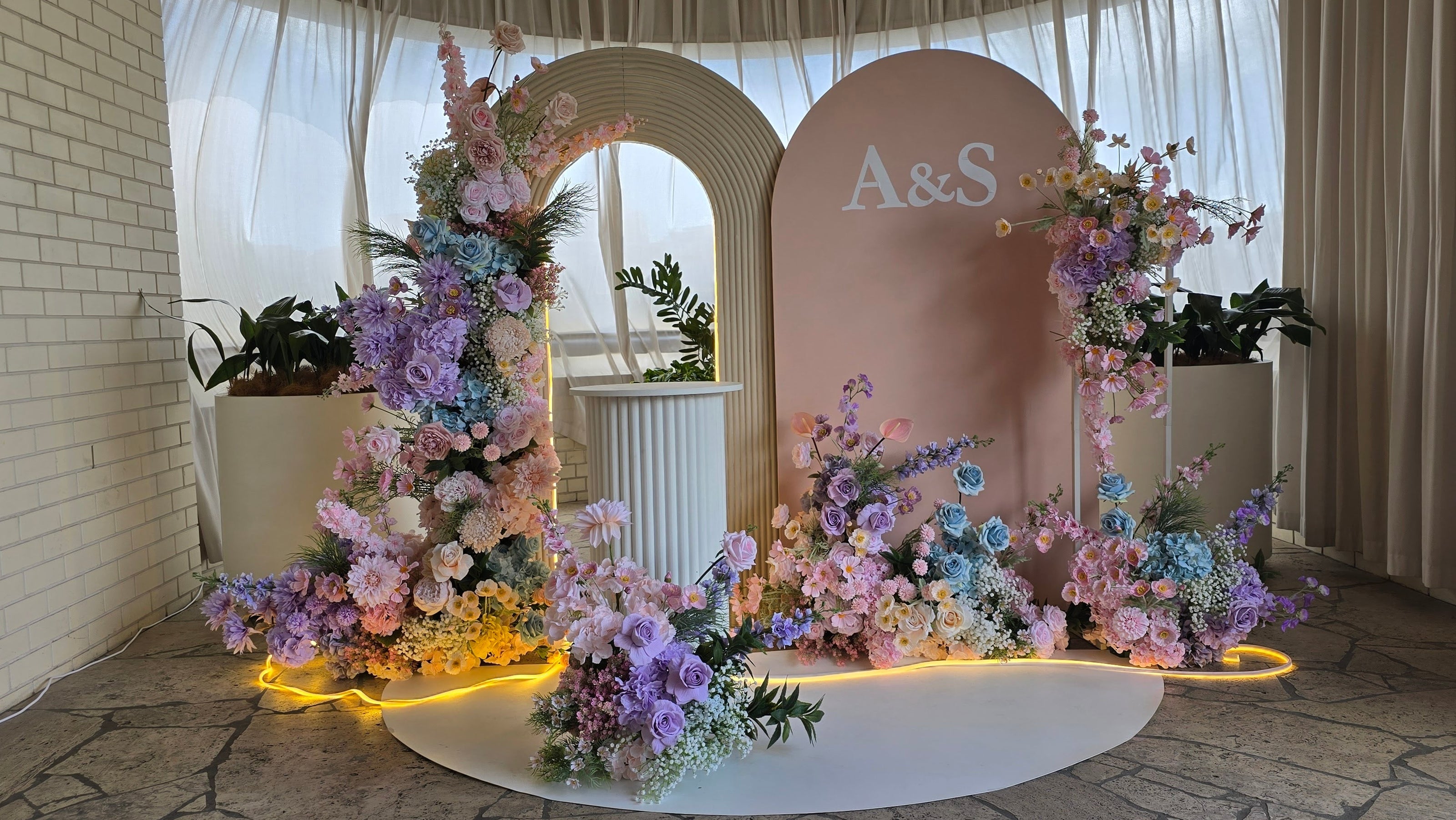 A styled wedding backdrop setup featuring a large pink arch with 'A&S' printed on it, flanked by floral arrangements in pastel purple, pink, and blue, with warm yellow accent lights on a stone floor. Perfect Styling for Weddings, engagements and receptions in Brisbane. Lush and Lavish events.