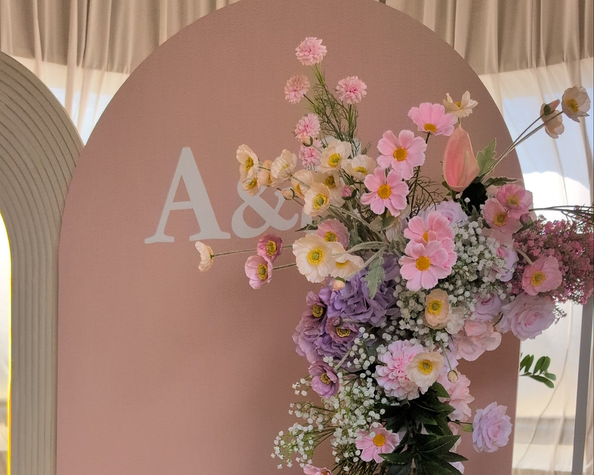 A decorative floral event backdrop with a large blush pink arch background, white monogram letters 'A&S' at the top, and cascading arrangements of pink, lavender, blue, and white flowers. A curved warm white floor light runs along the bottom edge. Lush and Lavish events at Calile Hotel, Brisbane, Australia.