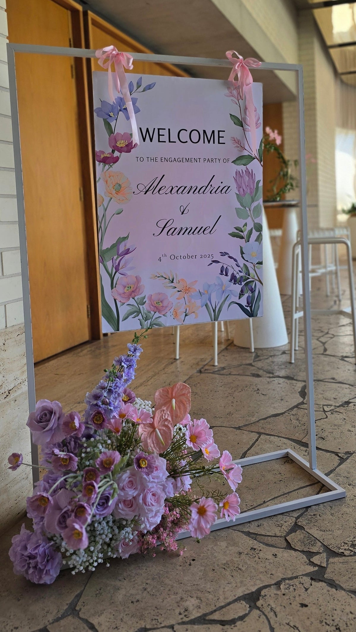 A wedding welcome sign on a metal stand in an outdoor patio setting, decorated with pink flowers and ribbons. The sign reads 'WELCOME to the engagement party of Alexandra & Samuel'. 