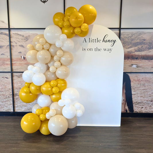 A balloon garland arrangement in yellow, cream, and white stands beside a white arch backdrop printed with the phrase 'A little honey is on the way.' The setup was created by Lush & Lavish Events a Brisbane and Gold Coast based Event planning, event styling and on the day coordination business.
