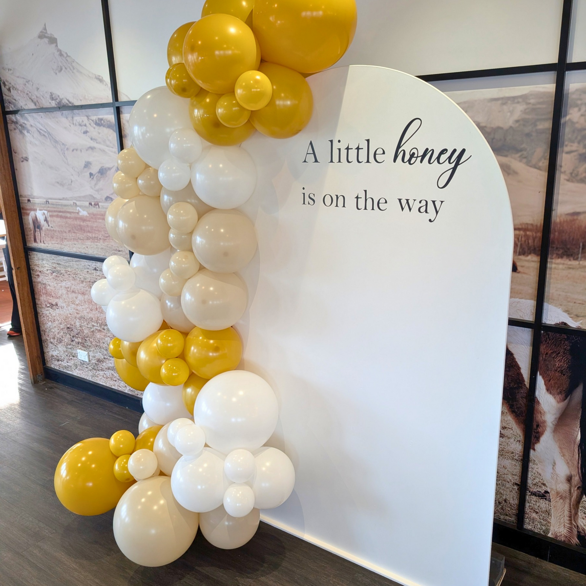 A white arched backdrop banner with the printed phrase 'A little honey is on the way' in black script, decorated on the left with a balloon garland in white, cream, and yellow tones. Backdrop created by Lush and Lavish Events a Brisbane and Gold Coast based event planning, event styling and on-the-day coordination business.