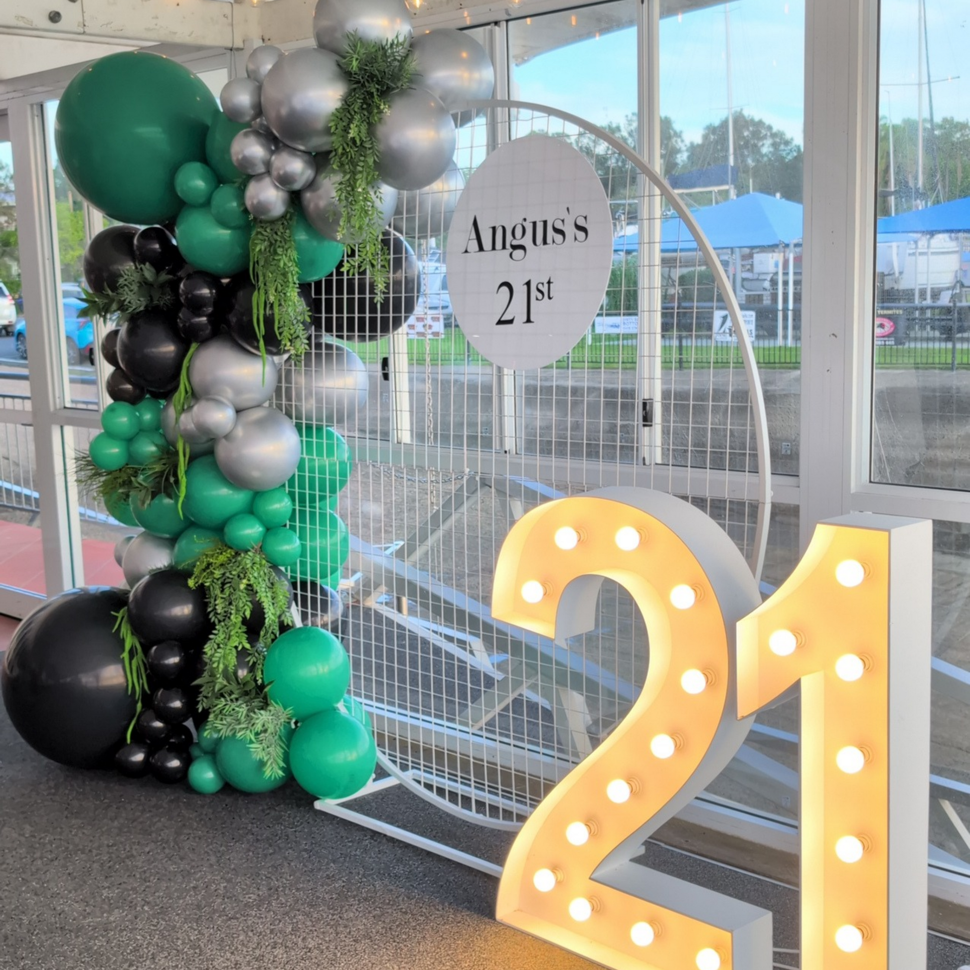 Lush green, silver and black 21st birthday balloon backdrop featuring a mesh arch, organic balloon garland with faux greenery accents and large illuminated LED number 21 lights. A luxury birthday party backdrop hire option perfect for 21st celebrations, milestone birthdays, photo opportunities and event styling across Brisbane, Gold Coast and South East Queensland