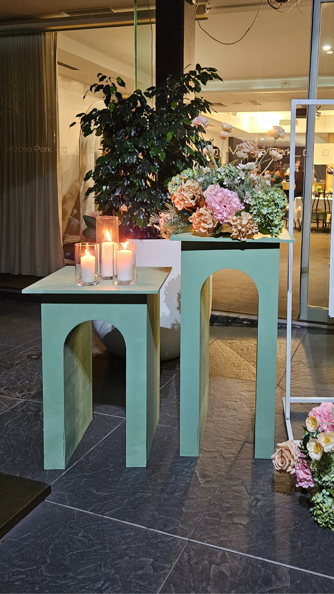 Two green plinths of different heights, each with an arch-shaped cutout in the middle. The shorter plinth holds three lit candles in glass holders, while the taller one is decorated with pink and white flowers. Prop hire, Prop hiring, Prop hire, event stylists Brisbane, brisbane event styling, brisbane event styling, event stylist Brisbane, Brisbane Wedding, Gold Coast Wedding, Floral Styling, Lush & Lavish Events