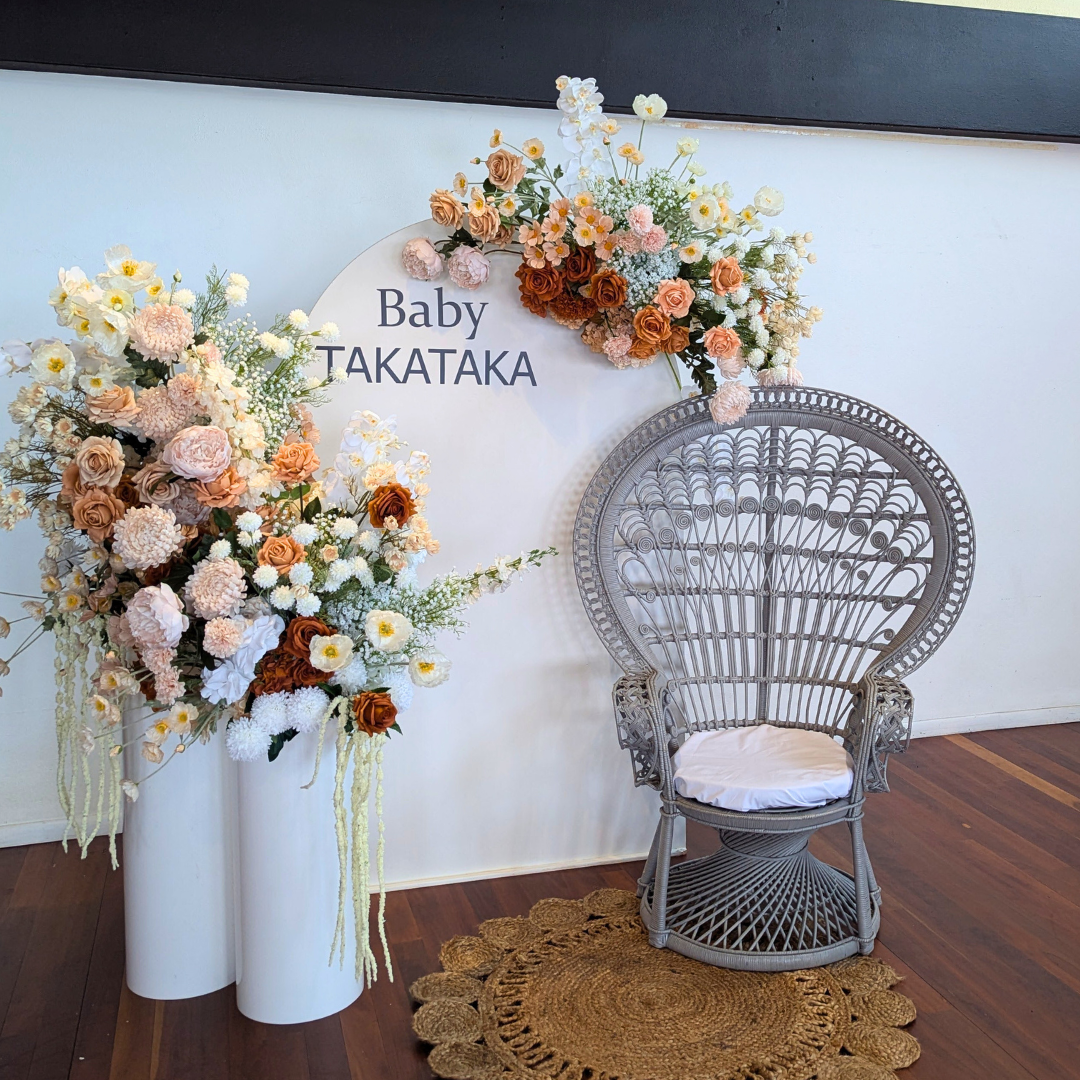 Decorative floral arrangement with 'Baby Takataka' text next to a wicker chair on a wooden floor. baby shower backdrop, neutral floral backdrop, elegant event styling, Brisbane party hire, floral arch setup, engagement backdrop, birthday backdrop hire, custom event decor, neutral baby shower theme, white arch floral arrangement