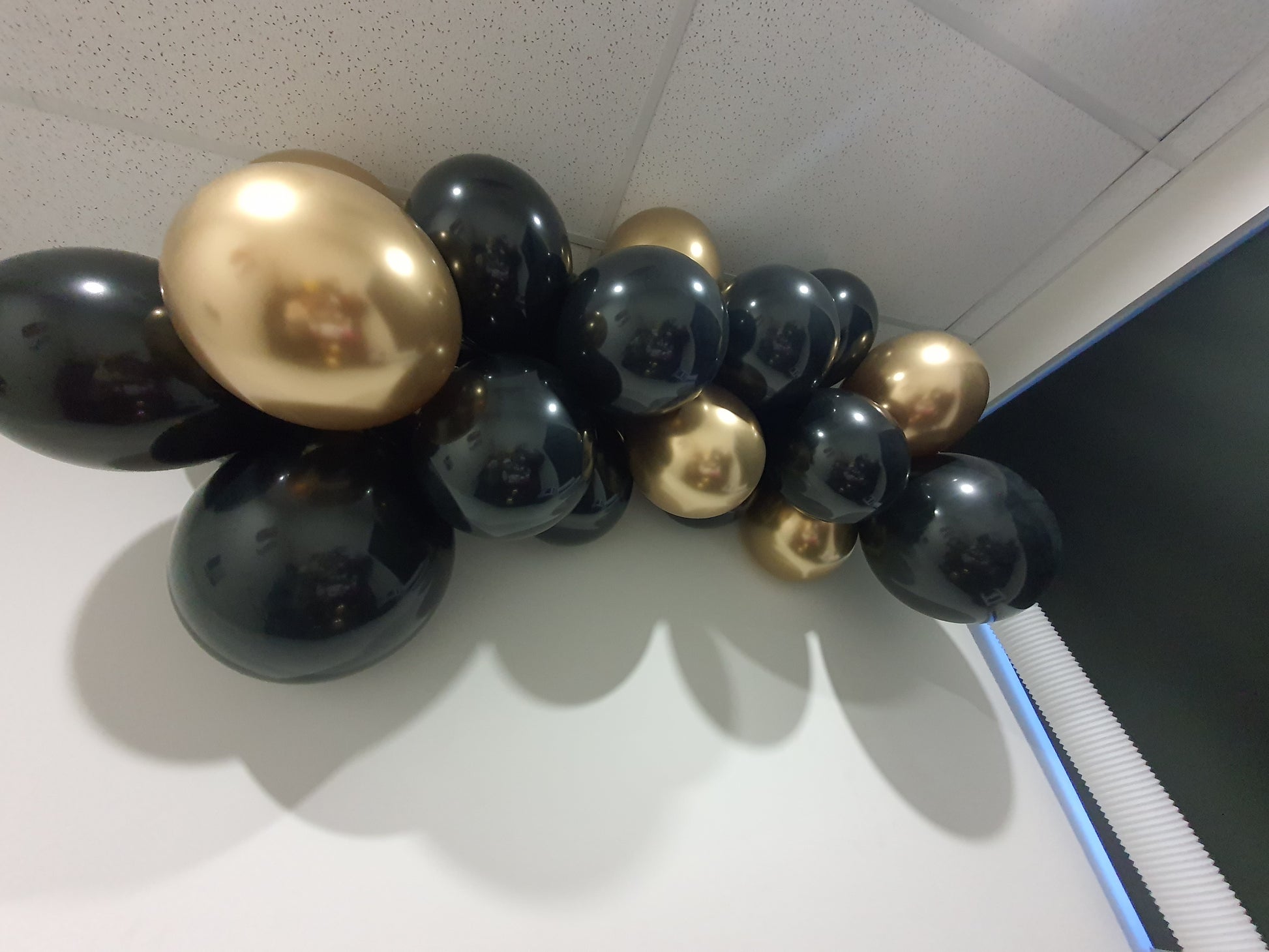 A cluster of black and gold balloons is attached to the ceiling corner of a room. The shiny, reflective balloons create a festive and elegant touch against a speckled ceiling. Prop hire, Prop hiring, Prop hire, event stylists Brisbane, brisbane event styling, brisbane event styling, event stylist Brisbane, Brisbane Wedding, Gold Coast Wedding, Floral Styling, Lush & Lavish Events, Brisbane Corporate Styling, Gold Coast Corporate Styling, Brisbane Brand Activation