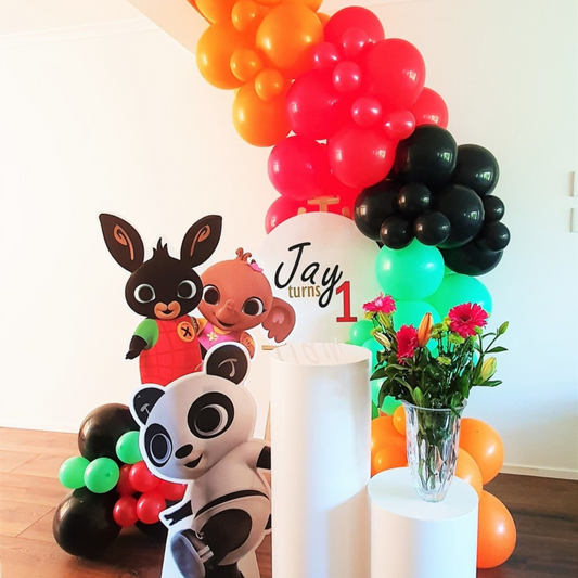 olorful first birthday party setup featuring a balloon arch in orange, black, and green colors, with life-size cutouts of cartoon characters including a panda and a rabbit. A white pedestal holds a vase with red flowers, and there’s a custom sign that reads ‘Jay turns 1’ for an eye-catching celebration backdrop. Prop hire, Prop hiring, Prop hire, event stylists Brisbane, brisbane event styling, brisbane event styling, event stylist Brisbane, Brisbane Wedding, Gold Coast Wedding, Floral Styling