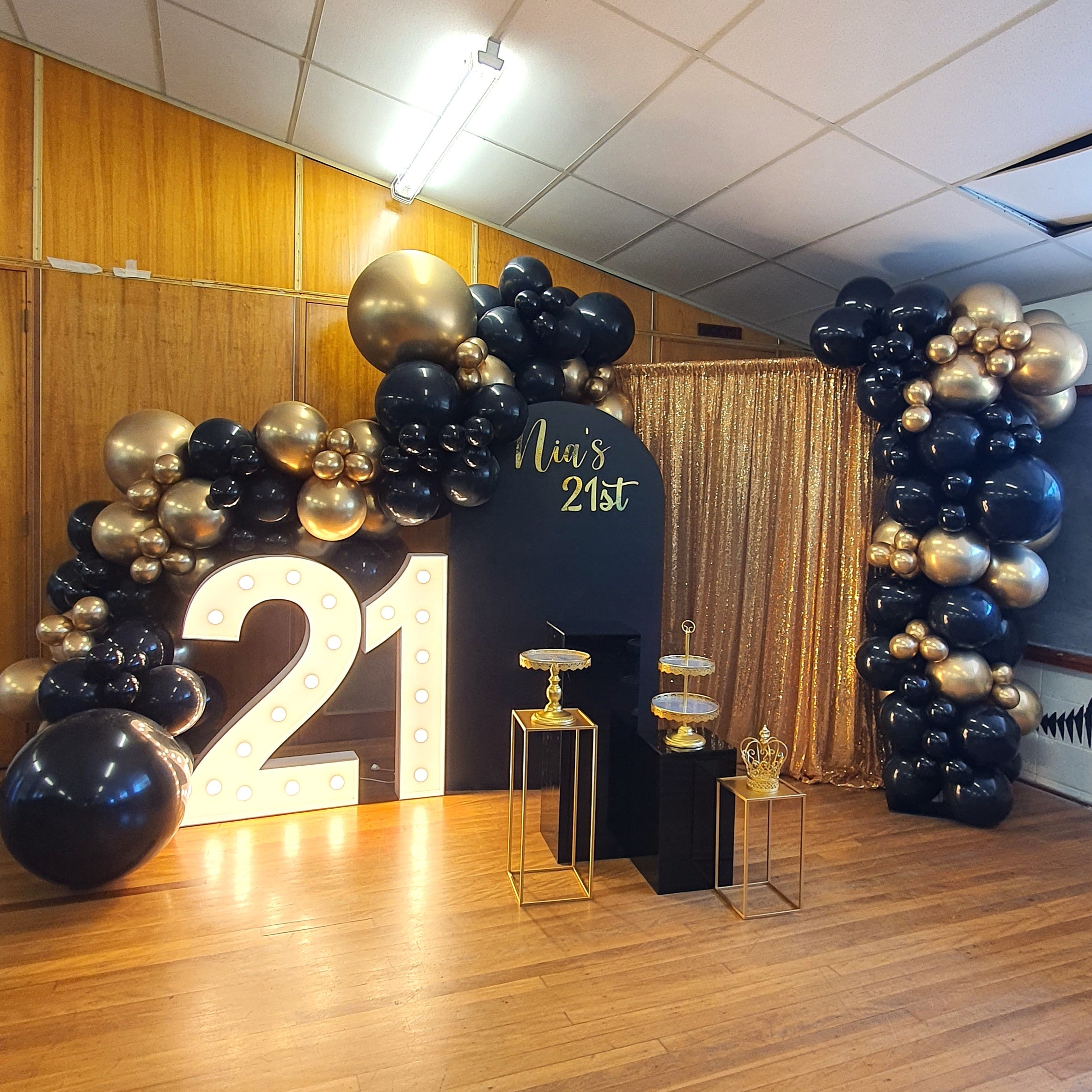 A decorated area for a 21st birthday celebration. The setup includes a large illuminated number "21" and a black backdrop with the text "Nia's 21st" in gold. The area is adorned with black and gold balloons arranged in arches on both sides. There are also gold and glass stands for displaying items, and a gold sequin curtain in the background. Prop hire, Prop hiring, Prop hire, event stylists Brisbane, brisbane event styling, brisbane event styling, event stylist Brisbane, Lush & Lavish Events
