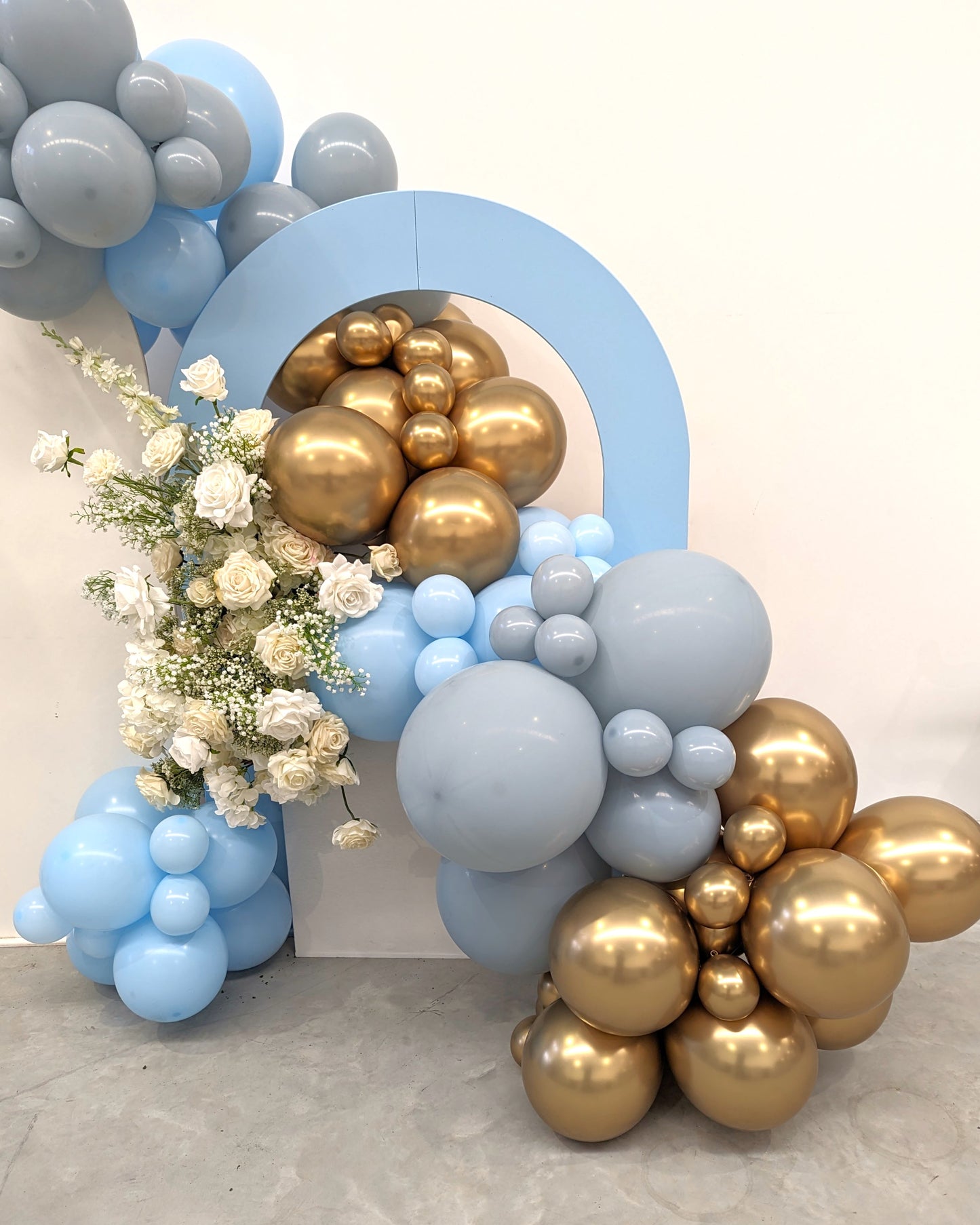 A decorative balloon arrangement featuring a mix of light blue, gray, and gold balloons. The balloons are arranged around a light blue arch structure. White flowers with green stems are interspersed among the balloons on the left side of the arrangement. Prop hire, Prop hiring, Prop hire, event stylists Brisbane, brisbane event styling, brisbane event styling, event stylist Brisbane, Brisbane Wedding, Gold Coast Wedding, Floral Styling, Lush & Lavish Events