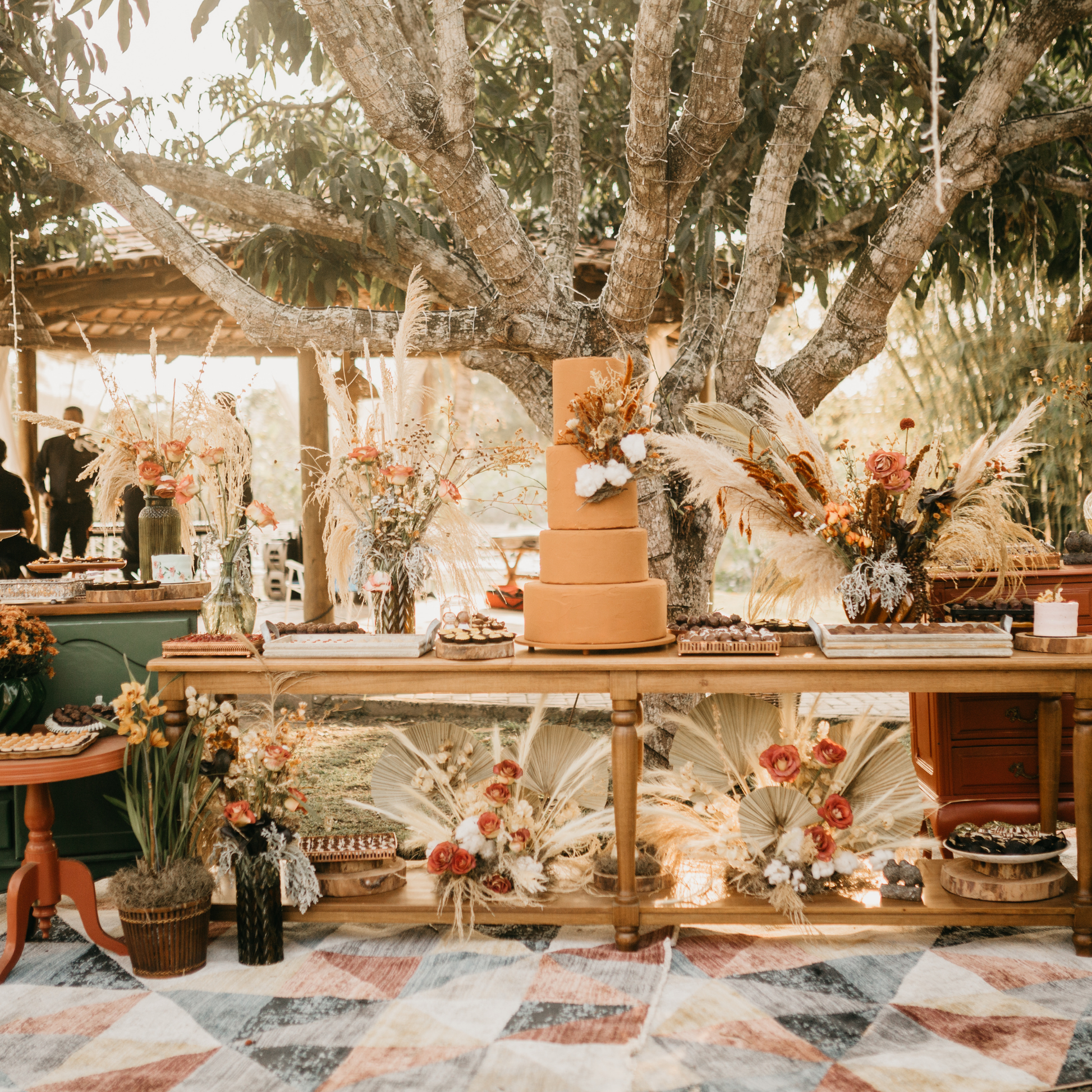 Rustic outdoor dessert table setup under a large tree with a multi-tiered cake, various desserts, and floral arrangements featuring dried flowers and pampas grass. A patterned blanket covers the ground, creating a bohemian atmosphere. Prop hire, Prop hiring, Prop hire, event stylists Brisbane, brisbane event styling, brisbane event styling, event stylist Brisbane, Brisbane Wedding, Gold Coast Wedding, Floral Styling, Lush & Lavish Events
