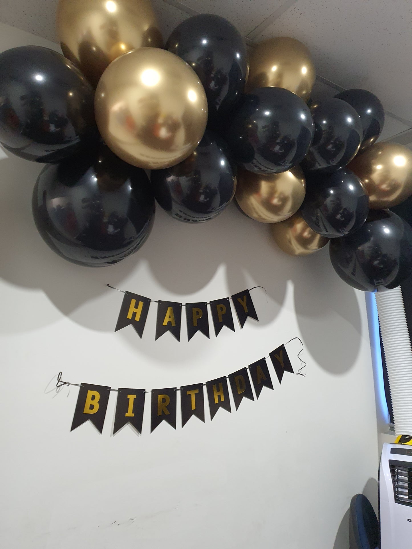 A wall decorated with black and gold balloons arranged in a cluster at the top. Below the balloons, there is a banner with black and gold flags spelling out "HAPPY BIRTHDAY." Prop hire, Prop hiring, Prop hire, event stylists Brisbane, brisbane event styling, brisbane event styling, event stylist Brisbane, Brisbane Wedding, Gold Coast Wedding, Floral Styling, Lush & Lavish Events, Brisbane Corporate Styling, Gold Coast Corporate Styling, Brisbane Brand Activation