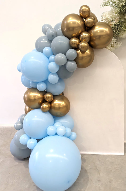Balloon arrangement with light blue, gray, and metallic gold balloons forming an arch, set against a white backdrop with greenery in the corner. Prop hire, Prop hiring, Prop hire, event stylists Brisbane, brisbane event styling, brisbane event styling, event stylist Brisbane, Brisbane Wedding, Gold Coast Wedding, Floral Styling, Lush & Lavish Events, Brisbane Tablescape, Gold Coast Tablescapes, Brisbane Candle Hire