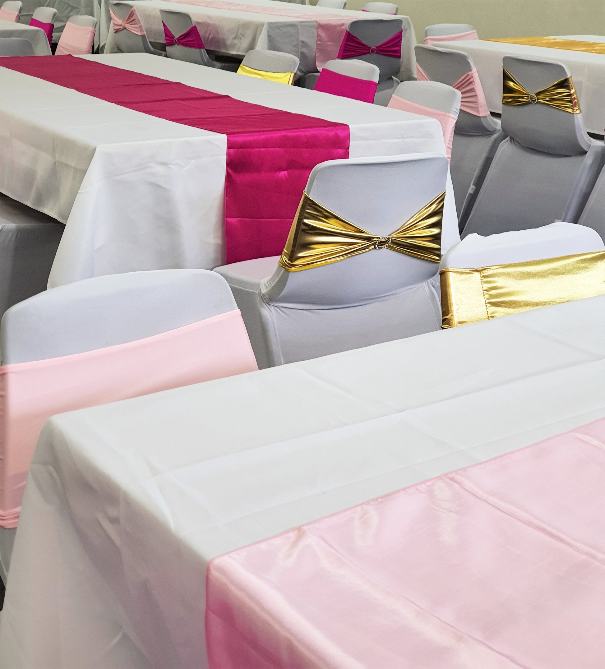 A decorated event space with tables covered in white cloths and colorful runners in shades of pink and red. Chairs are adorned with gray covers and sashes in gold, pink, and red, suggesting a festive or formal gathering. Prop hire, Prop hiring, Prop hire, event stylists Brisbane, brisbane event styling, brisbane event styling, event stylist Brisbane, Brisbane Wedding, Gold Coast Wedding, Floral Styling, Lush & Lavish Events