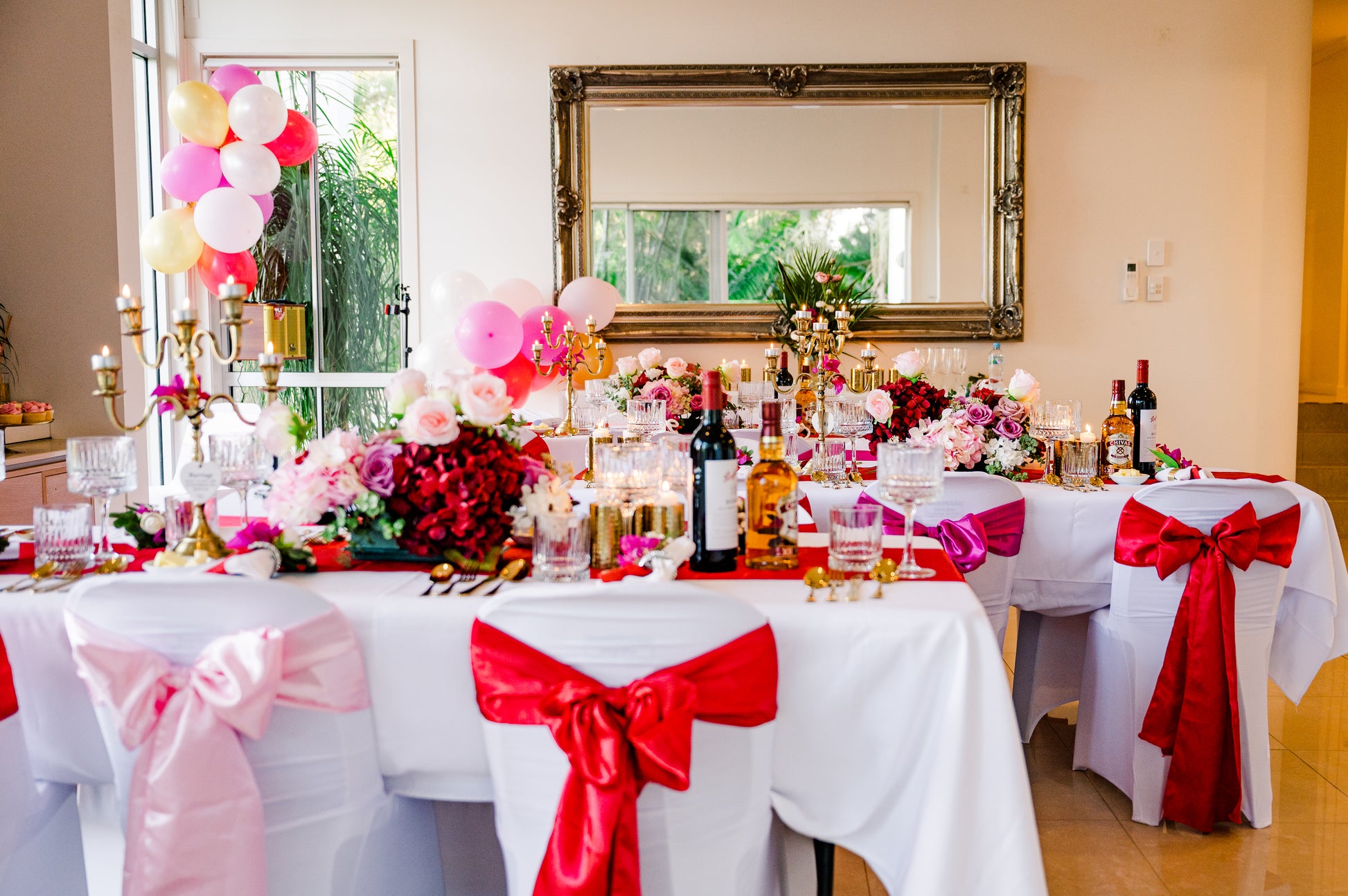A decorated dining table with pink and red ribbons, floral arrangements, candles, and bottles of wine and spirits. Balloons in pink, white, and gold colors are part of the festive setup. A large mirror hangs on the wall in the background. Prop hire, Prop hiring, Prop hire, event stylists Brisbane, brisbane event styling, brisbane event styling, event stylist Brisbane, Brisbane Wedding, Gold Coast Wedding, Floral Styling, Lush & Lavish Events