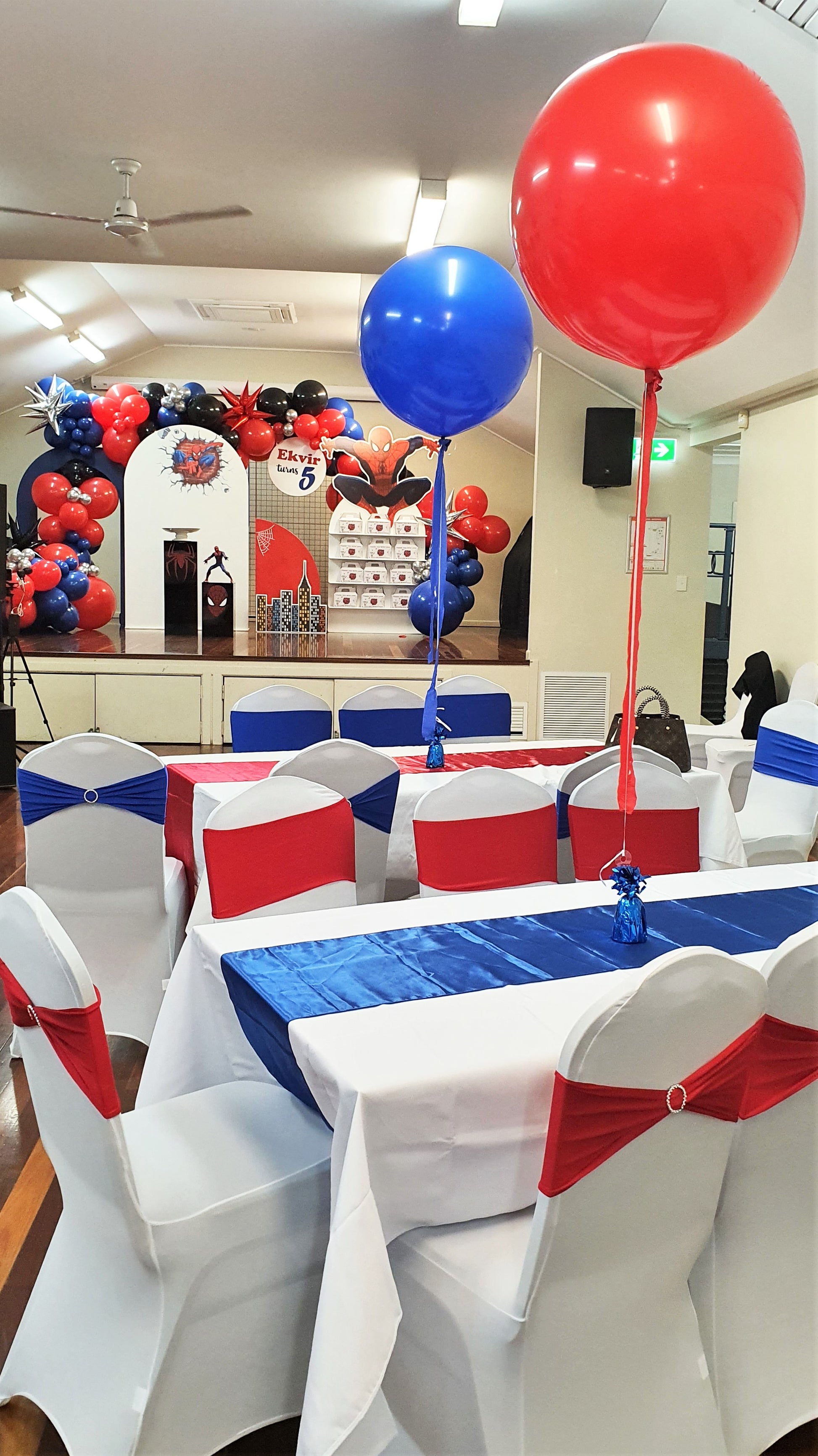 A decorated event hall with tables and chairs covered in white cloth. Chairs have red and blue sashes. Tables have blue runners with large red and blue balloons tied to them. A stage with a balloon arch and "Happy 5th Birthday" sign is in the background. Prop hire, Prop hiring, Prop hire, event stylists Brisbane, brisbane event styling, brisbane event styling, event stylist Brisbane, Brisbane Wedding, Gold Coast Wedding, Floral Styling, Lush & Lavish Events