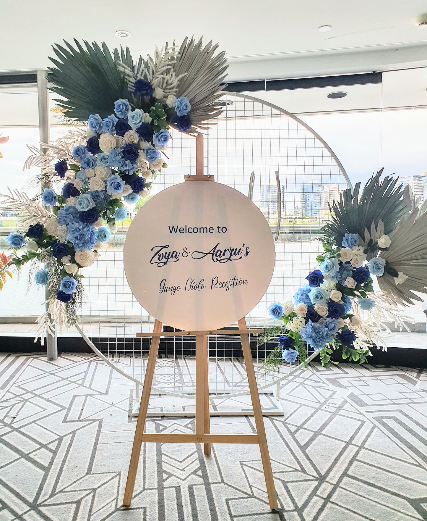 Circular welcome sign on an easel, reading "Welcome to Priya & Aarav's Sangeet & Reception," surrounded by blue and white flowers and dried palm leaves. A cityscape is visible through a large window in the background. Prop hire, Prop hiring, Prop hire, event stylists Brisbane, brisbane event styling, brisbane event styling, event stylist Brisbane, Brisbane Wedding, Gold Coast Wedding, Floral Styling, Lush & Lavish Events