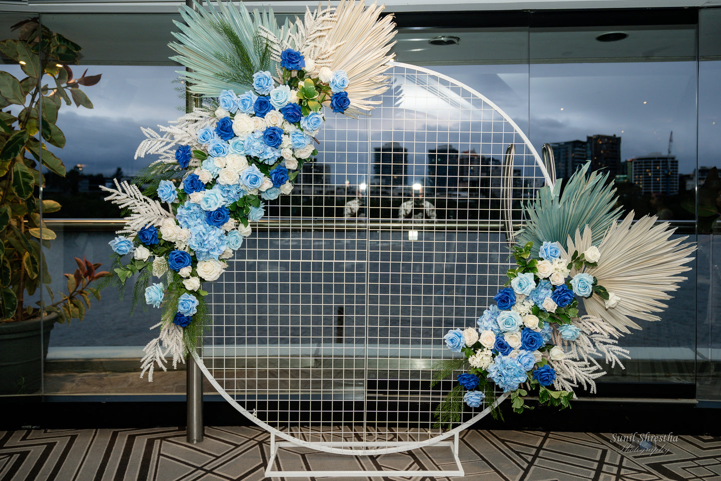 A decorative circular metal frame adorned with blue, white, and light green flowers, along with white and green foliage. The frame features a geometric grid pattern in the center. Prop hire, Prop hiring, Prop hire, event stylists Brisbane, brisbane event styling, brisbane event styling, event stylist Brisbane, Brisbane Wedding, Gold Coast Wedding, Floral Styling, Lush & Lavish Events