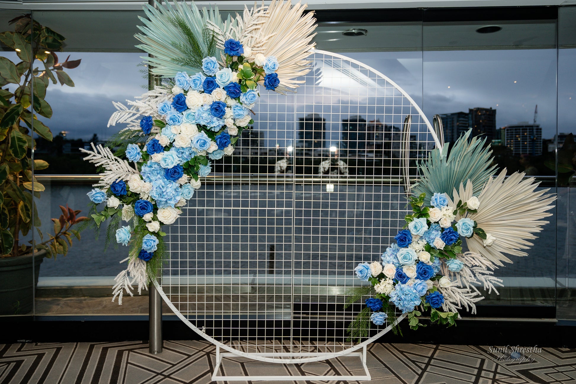 A decorative circular metal frame adorned with blue, white, and light green flowers, along with white and green foliage. The frame features a geometric grid pattern in the center. Prop hire, Prop hiring, Prop hire, event stylists Brisbane, brisbane event styling, brisbane event styling, event stylist Brisbane, Brisbane Wedding, Gold Coast Wedding, Floral Styling, Lush & Lavish Events