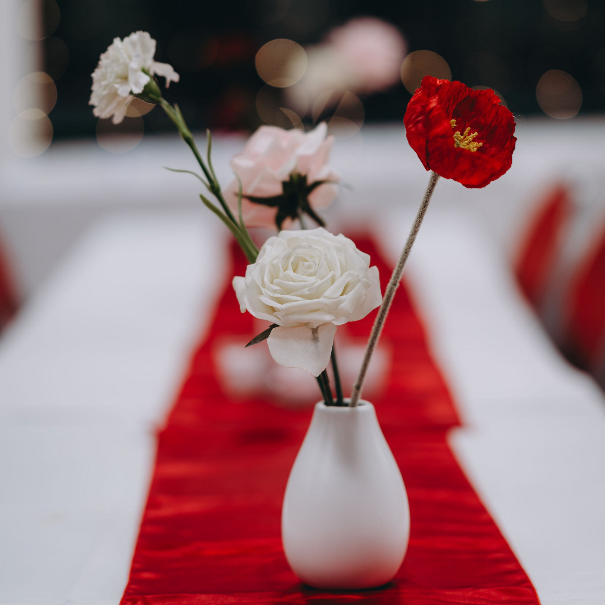 A white vase sits on a red table runner. Inside, a white rose, a pink rose, a red carnation, and a white carnation are arranged in a simple and elegant display. Prop hire, Prop hiring, Prop hire, event stylists Brisbane, brisbane event styling, brisbane event styling, event stylist Brisbane, Brisbane Wedding, Gold Coast Wedding, Floral Styling, Lush & Lavish Events, Brisbane Tablescape, Gold Coast Tablescapes, Brisbane Candle Hire