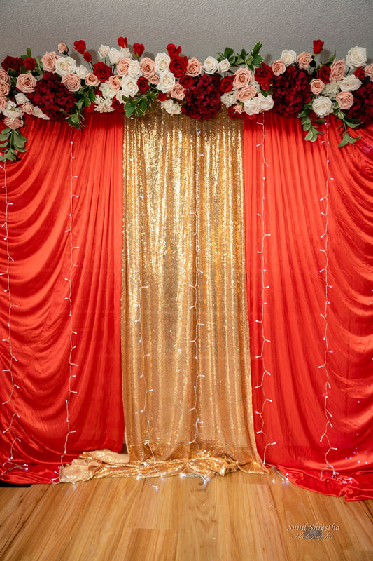 Decorative backdrop with red and gold draped fabric, a gold sparkly center panel, and floral arrangements of roses. String lights hang vertically along the red fabric, enhancing the festive atmosphere. Prop hire, Prop hiring, Prop hire, event stylists Brisbane, brisbane event styling, brisbane event styling, event stylist Brisbane, Brisbane Wedding, Gold Coast Wedding, Floral Styling, Lush & Lavish Events