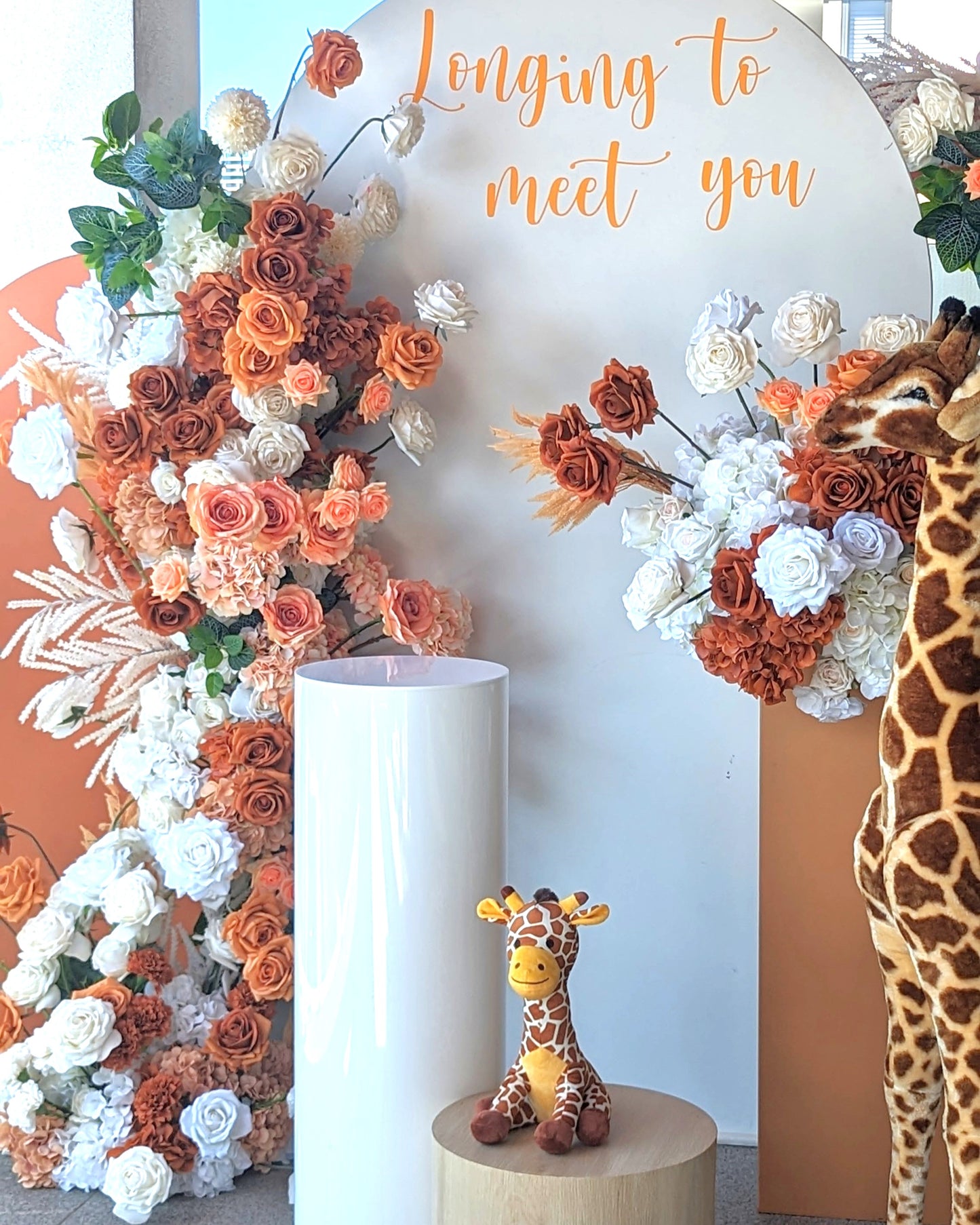 Two large giraffe statues and one small giraffe statue on a wooden stool. The backdrop includes a white and orange arch with orange and white flowers, with the text “longing to meet you.” Prop hire, Prop hiring, Prop hire, event stylists Brisbane, brisbane event styling, brisbane event styling, event stylist Brisbane, Brisbane Wedding, Gold Coast Wedding, Floral Styling, Lush & Lavish Events, Brisbane Tablescape, Gold Coast Tablescapes, Brisbane Candle Hire