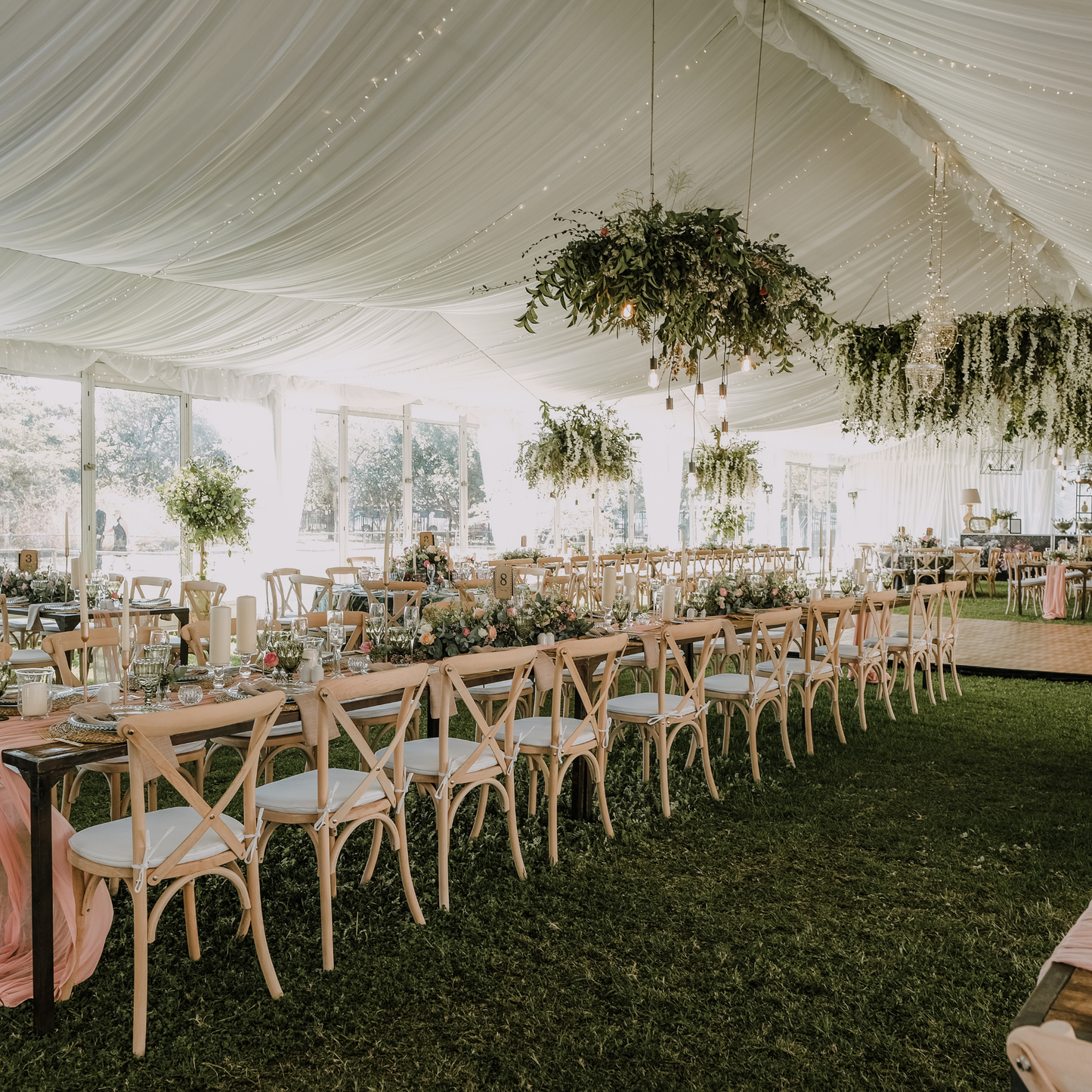 A wedding reception is held under a large white tent adorned with greenery and hanging lights. Tables with white tablecloths and cross-back chairs are set up on a lush green lawn. Prop hire, Prop hiring, Prop hire, event stylists Brisbane, brisbane event styling, brisbane event styling, event stylist Brisbane, Brisbane Wedding, Gold Coast Wedding, Floral Styling, Lush & Lavish Events, Brisbane Tablescape, Gold Coast Tablescapes, Brisbane Candle Hire