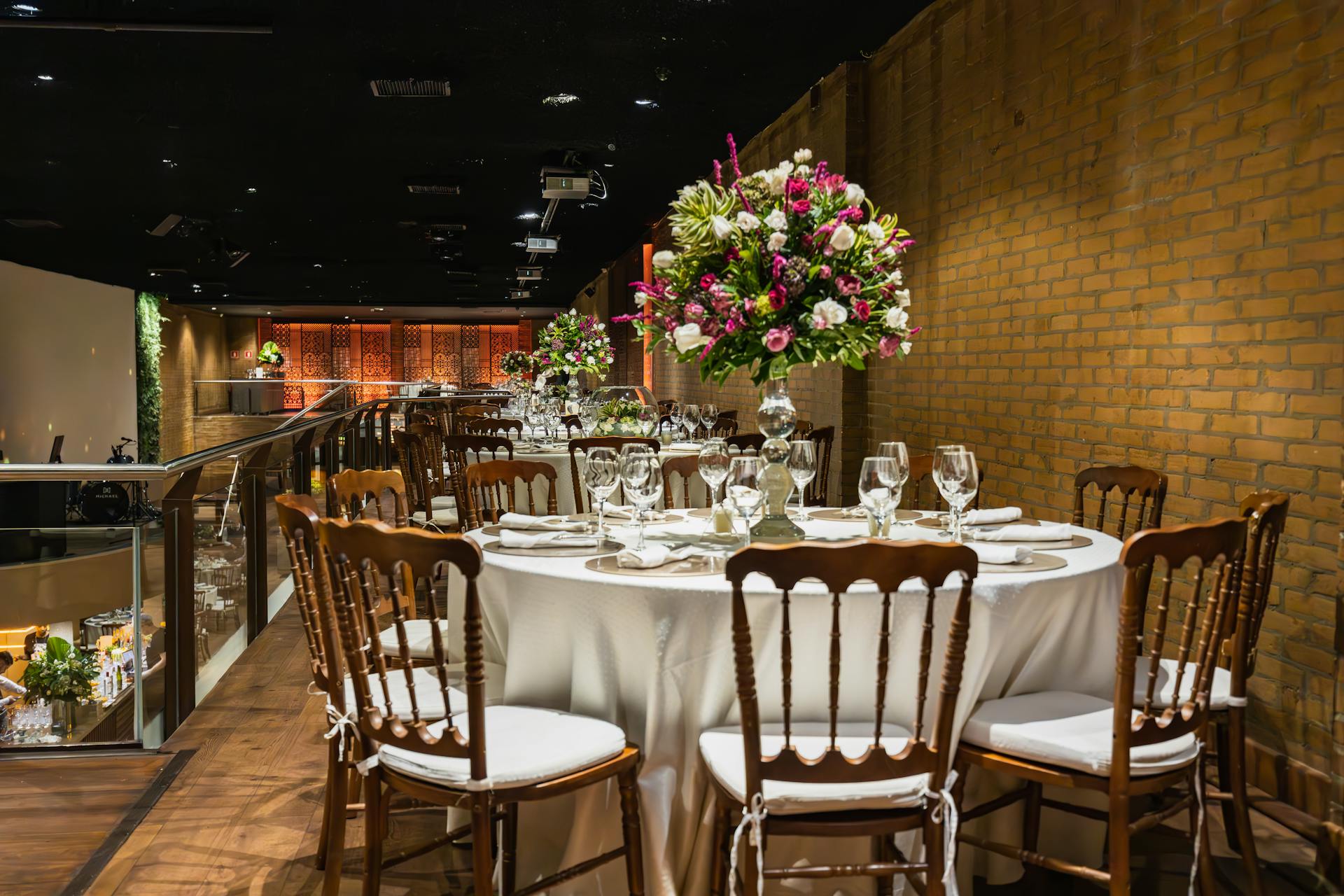 A round dining table with a white tablecloth and floral centerpiece, surrounded by wooden chairs with white cushions. The setting is in an elegant, dimly lit restaurant or banquet hall with brick walls and a wooden floor.  Prop hire, Prop hiring, Prop hire, event stylists Brisbane, brisbane event styling, brisbane event styling, event stylist Brisbane, Brisbane Wedding, Gold Coast Wedding, Floral Styling, Lush & Lavish Events