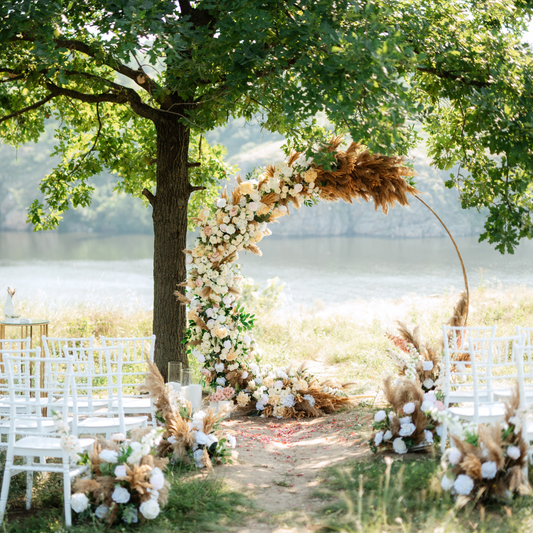 Outdoor wedding under a tree by a lake. Floral arch with white and beige flowers and pampas grass. White chairs line an aisle with more floral arrangements. Serene lake and greenery in the background. Prop hire, Prop hiring, Prop hire, event stylists Brisbane, brisbane event styling, brisbane event styling, event stylist Brisbane, Brisbane Wedding, Gold Coast Wedding, Floral Styling, Lush & Lavish Events