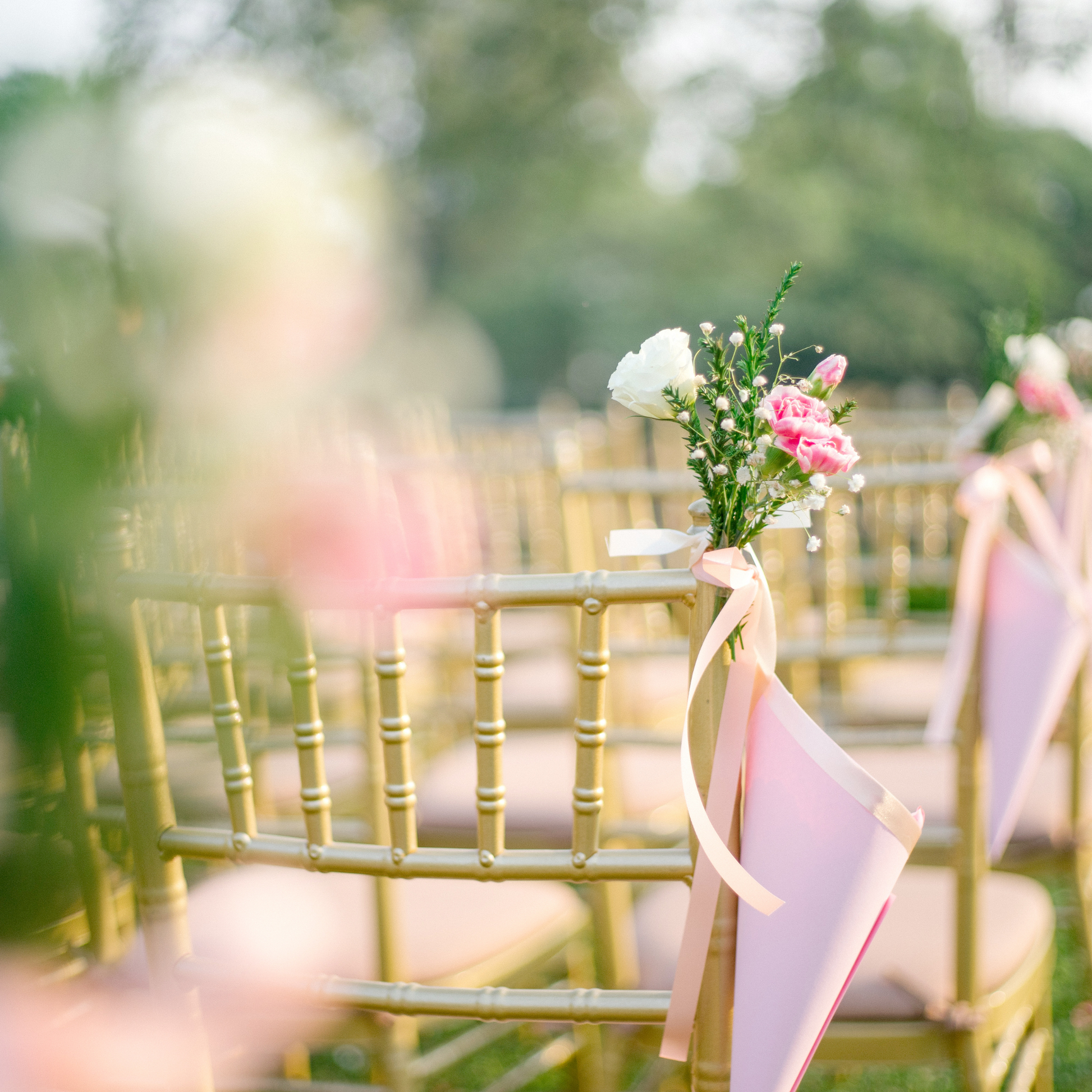 Rows of gold-colored chairs with small bouquets of pink and white flowers tied with pink ribbons, set up outdoors for an event. Prop hire, Prop hiring, Prop hire, event stylists Brisbane, brisbane event styling, brisbane event styling, event stylist Brisbane, Brisbane Wedding, Gold Coast Wedding, Floral Styling, Lush & Lavish Events