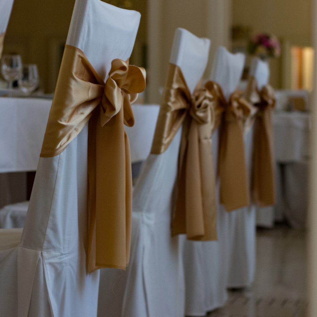 A row of white chairs with gold bows tied around the backrests, arranged in a formal setting, likely for an event such as a wedding or banquet. The chairs are aligned neatly in front of a table set with glassware. Prop hire, Prop hiring, Prop hire, event stylists Brisbane, brisbane event styling, brisbane event styling, event stylist Brisbane, Brisbane Wedding, Gold Coast Wedding, Floral Styling, Lush & Lavish Events