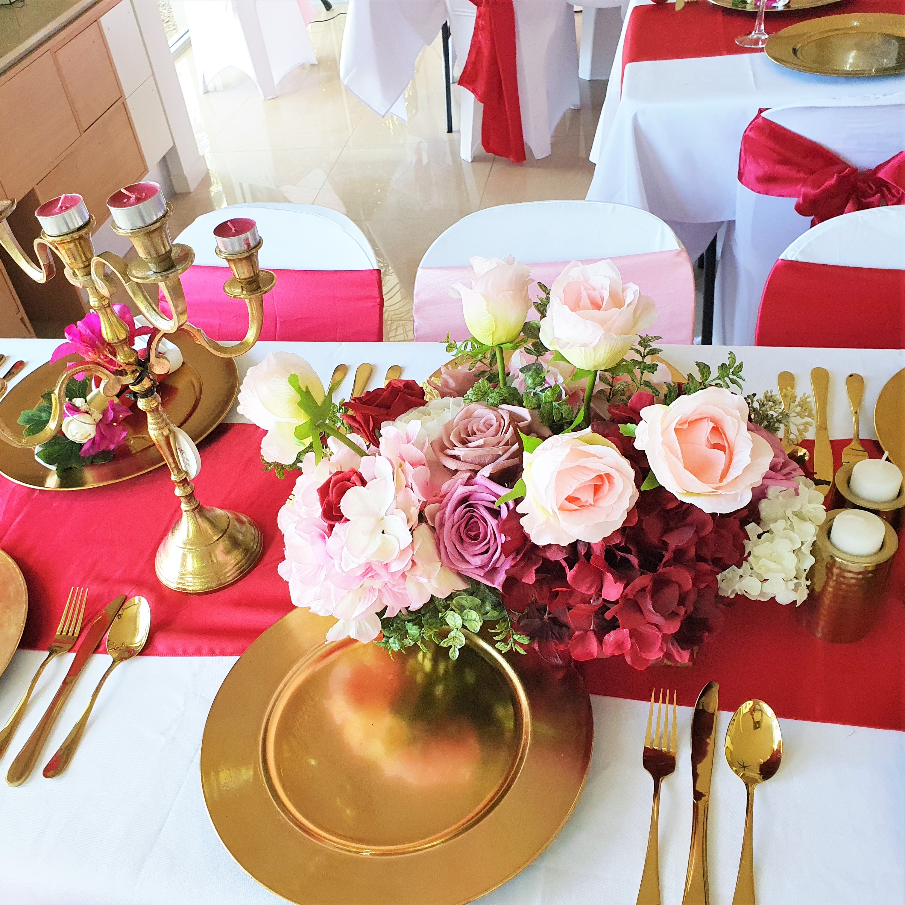 A festive table setting features a centerpiece of pink and red roses, gold chargers, red napkins, and gold cutlery. A gold candelabra with lit candles completes the elegant display. Prop hire, Prop hiring, Prop hire, event stylists Brisbane, brisbane event styling, brisbane event styling, event stylist Brisbane, Brisbane Wedding, Gold Coast Wedding, Floral Styling, Lush & Lavish Events, Brisbane Tablescape, Gold Coast Tablescapes, Brisbane Candle Hire