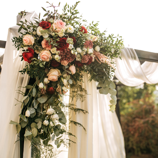 A floral arrangement featuring a mix of red, pink, and white roses, along with other greenery and flowers, is draped over a wooden arch adorned with white fabric. The arrangement is lush and cascading, creating an elegant and romantic atmosphere, likely for a wedding or special event. The background shows a blurred outdoor setting with greenery. Prop hire, Prop hiring, Prop hire, event stylists Brisbane, brisbane event styling, brisbane event styling, event stylist Brisbane, Brisbane Wedding, GC Wedding