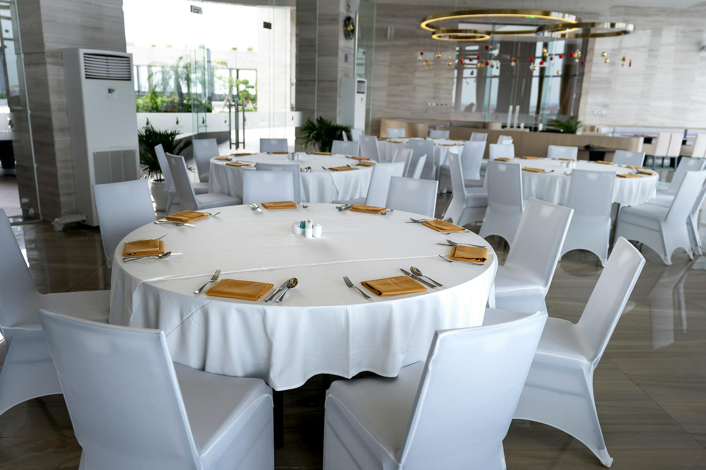 Modern dining area with round tables in white tablecloths, yellow napkins, and white chairs, featuring large windows and a circular light fixture. Prop hire, Prop hiring, Prop hire, event stylists Brisbane, brisbane event styling, brisbane event styling, event stylist Brisbane, Brisbane Wedding, Gold Coast Wedding, Floral Styling, Lush & Lavish Events