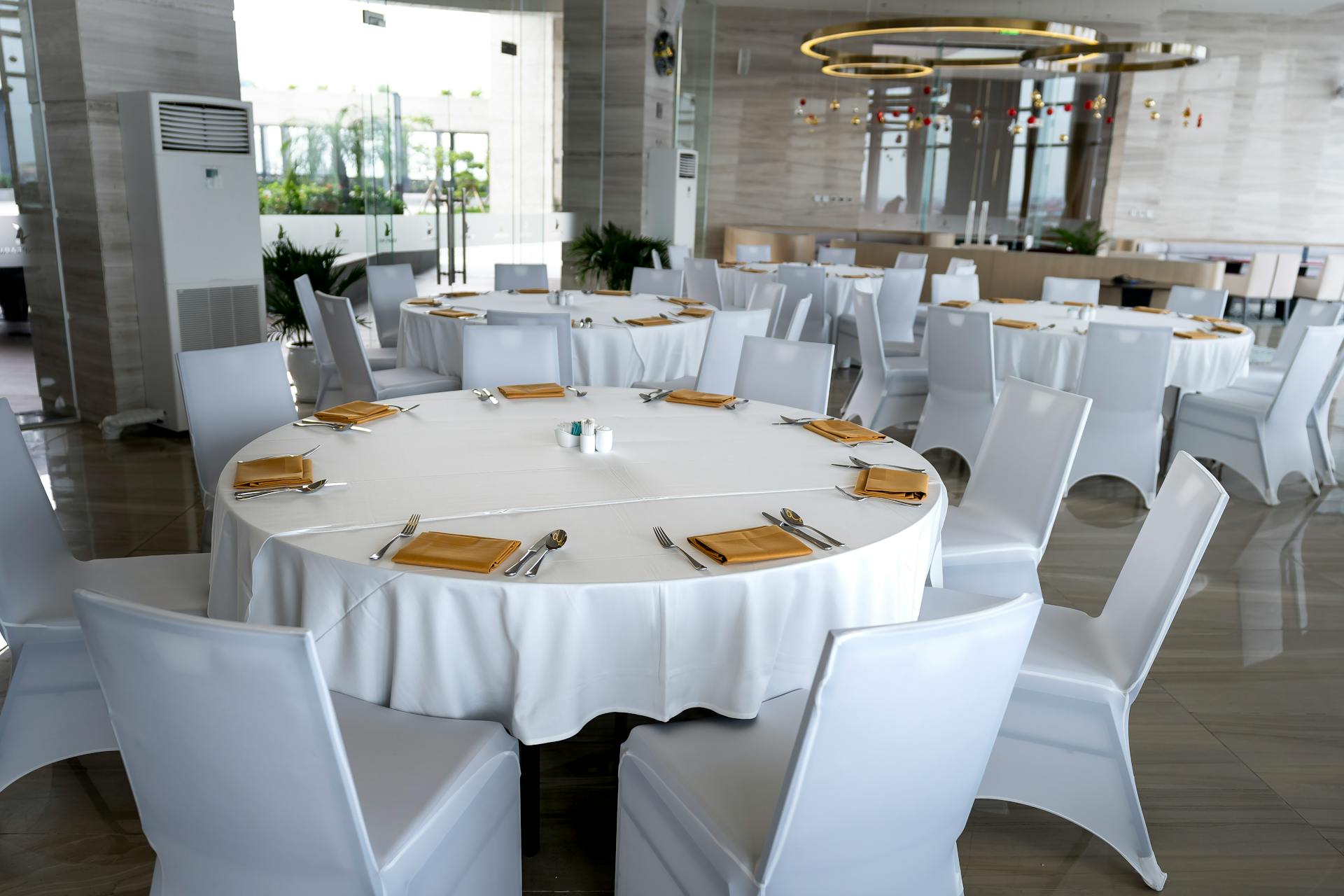 Modern dining area with round tables in white tablecloths, yellow napkins, and white chairs, featuring large windows and a circular light fixture. Prop hire, Prop hiring, Prop hire, event stylists Brisbane, brisbane event styling, brisbane event styling, event stylist Brisbane, Brisbane Wedding, Gold Coast Wedding, Floral Styling, Lush & Lavish Events