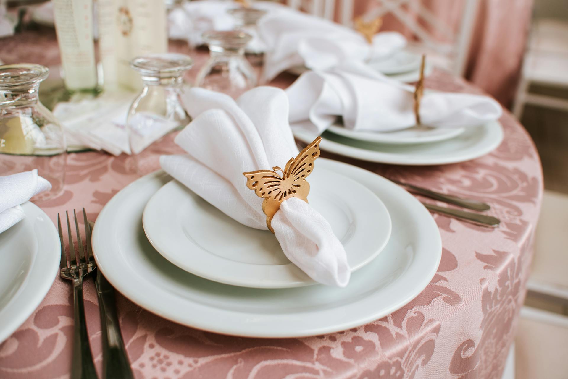A table is set with white plates and napkins. Each napkin is folded and held in place with a gold butterfly napkin ring. The table is covered with a pink tablecloth. Prop hire, Prop hiring, Prop hire, event stylists Brisbane, brisbane event styling, brisbane event styling, event stylist Brisbane, Brisbane Wedding, Gold Coast Wedding, Floral Styling, Lush & Lavish Events, Brisbane Tablescape, Gold Coast Tablescapes, Brisbane Candle Hire