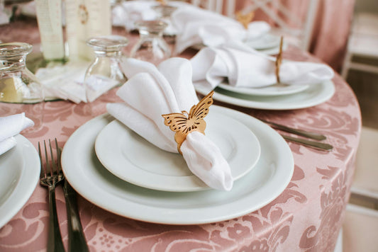 A table is set with white plates and napkins. Each napkin is folded and held in place with a gold butterfly napkin ring. The table is covered with a pink tablecloth. Prop hire, Prop hiring, Prop hire, event stylists Brisbane, brisbane event styling, brisbane event styling, event stylist Brisbane, Brisbane Wedding, Gold Coast Wedding, Floral Styling, Lush & Lavish Events, Brisbane Tablescape, Gold Coast Tablescapes, Brisbane Candle Hire