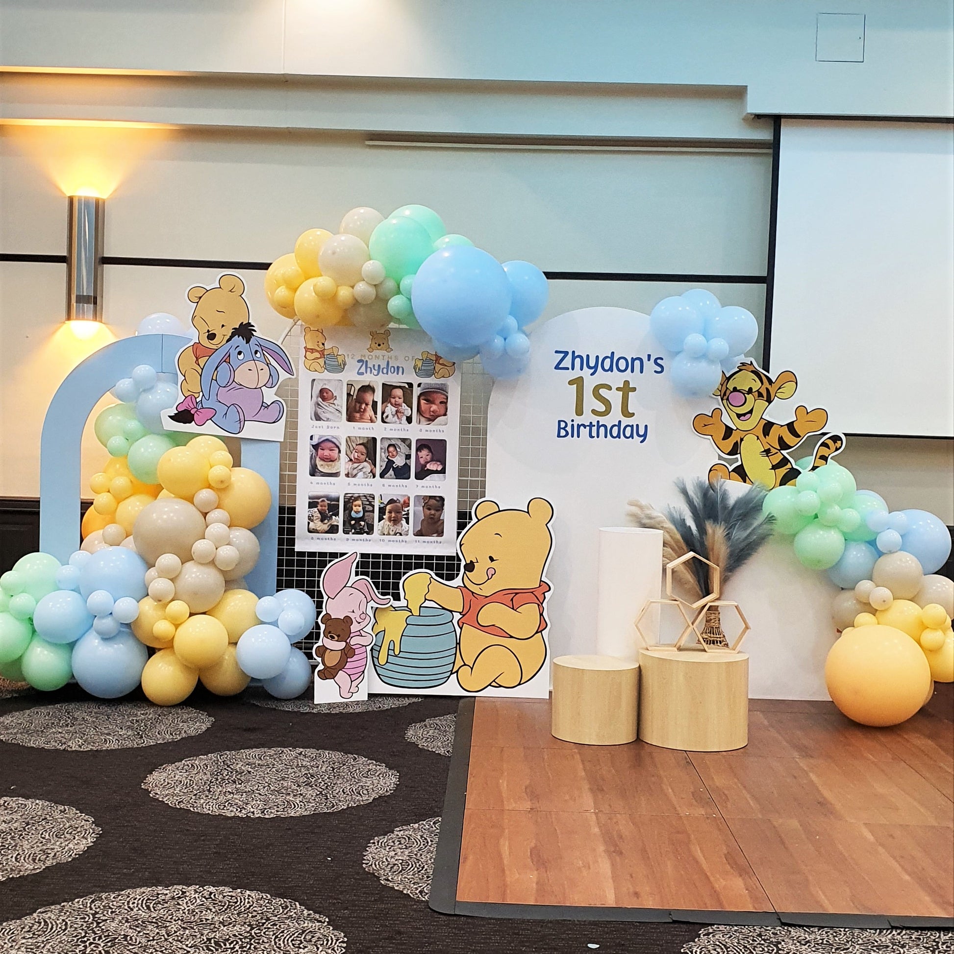 A Winnie the Pooh themed first birthday party backdrop features a balloon arch in blue, yellow, and white. Cutouts of Winnie the Pooh characters, along with a photo collage and wooden blocks. Prop hire, Prop hiring, Prop hire, event stylists Brisbane, brisbane event styling, brisbane event styling, event stylist Brisbane, Brisbane Wedding, Gold Coast Wedding, Floral Styling, Lush & Lavish Events, Brisbane Tablescape, Gold Coast Tablescapes, Brisbane Candle Hire