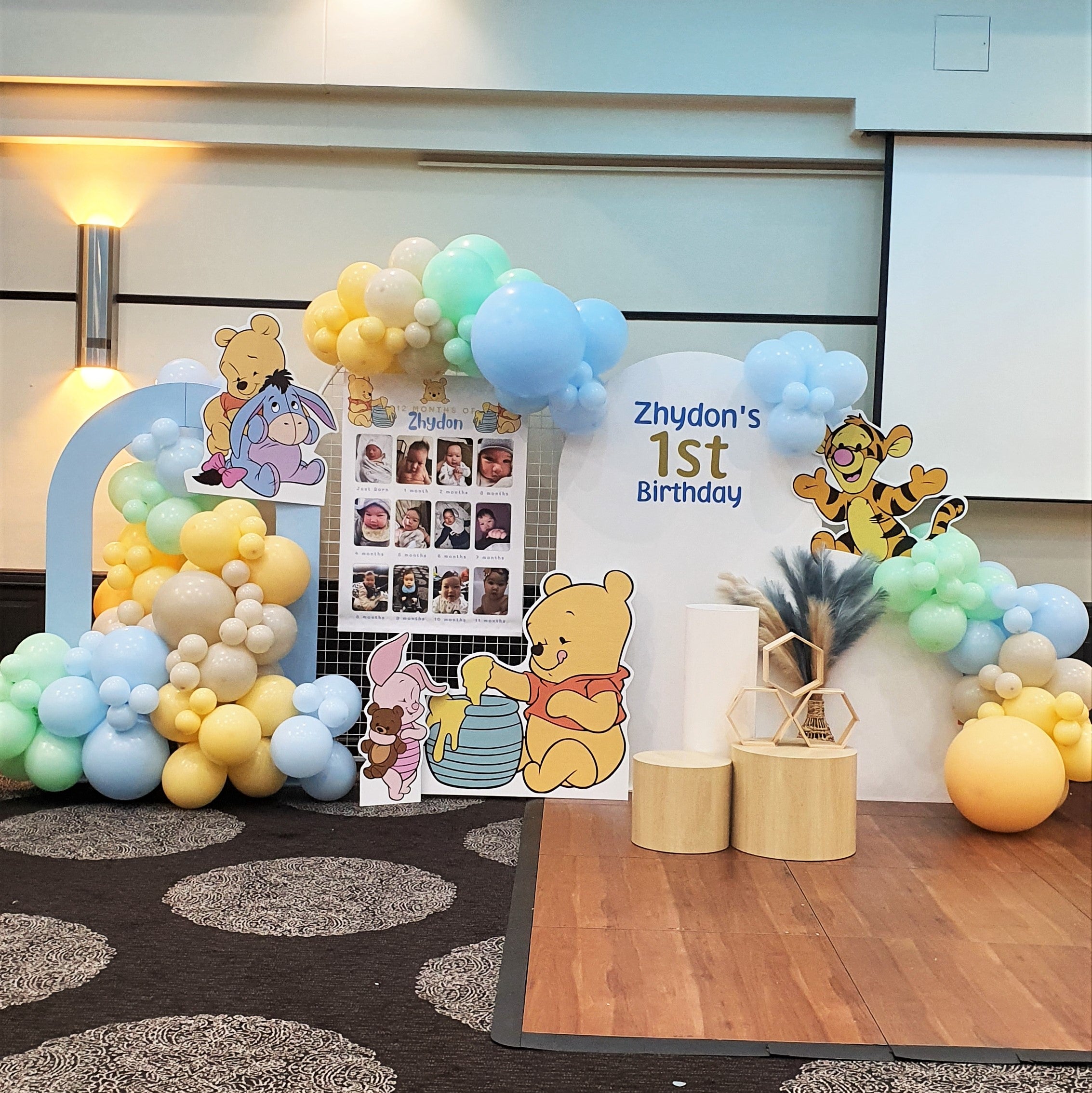A Winnie the Pooh themed first birthday party backdrop features a balloon arch in blue, yellow, and white. Cutouts of Winnie the Pooh characters, along with a photo collage and wooden blocks. Prop hire, Prop hiring, Prop hire, event stylists Brisbane, brisbane event styling, brisbane event styling, event stylist Brisbane, Brisbane Wedding, Gold Coast Wedding, Floral Styling, Lush & Lavish Events, Brisbane Tablescape, Gold Coast Tablescapes, Brisbane Candle Hire