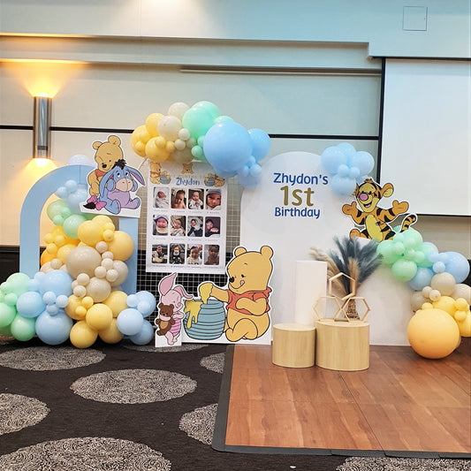 A Winnie the Pooh themed first birthday party backdrop features a balloon arch in blue, yellow, and white. Cutouts of Winnie the Pooh characters, along with a photo collage and wooden blocks. Prop hire, Prop hiring, Prop hire, event stylists Brisbane, brisbane event styling, brisbane event styling, event stylist Brisbane, Brisbane Wedding, Gold Coast Wedding, Floral Styling, Lush & Lavish Events, Brisbane Tablescape, Gold Coast Tablescapes, Brisbane Candle Hire