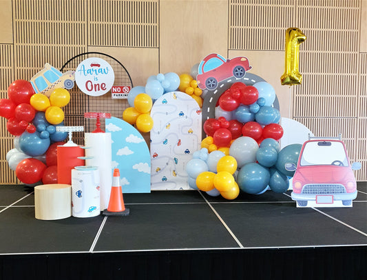 A colorful birthday party setup with a car theme. Backdrop features cars and clouds, balloon clusters in red, blue, yellow, and gray. A sign reads "Aarav is One" with a "No Parking" sign below. A large gold balloon shaped like the number 1 is on the right, with props like a traffic cone and cylindrical stands in front. Prop hire, Prop hiring, Prop hire, event stylists Brisbane, brisbane event styling, brisbane event styling, event stylist Brisbane, Brisbane Wedding, Gold Coast Wedding, Floral Styling