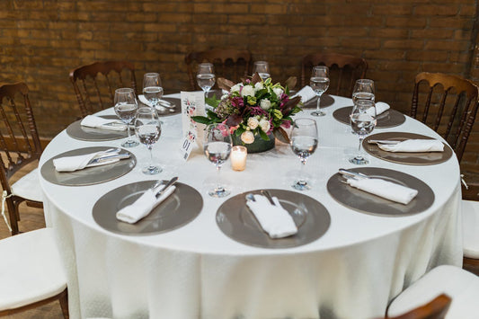 Round table set for a formal dinner with eight place settings, a floral centerpiece, and a lit candle. The table has a white tablecloth and is surrounded by wooden chairs. Prop hire, Prop hiring, Prop hire, event stylists Brisbane, brisbane event styling, brisbane event styling, event stylist Brisbane, Brisbane Wedding, Gold Coast Wedding, Floral Styling, Lush & Lavish Events