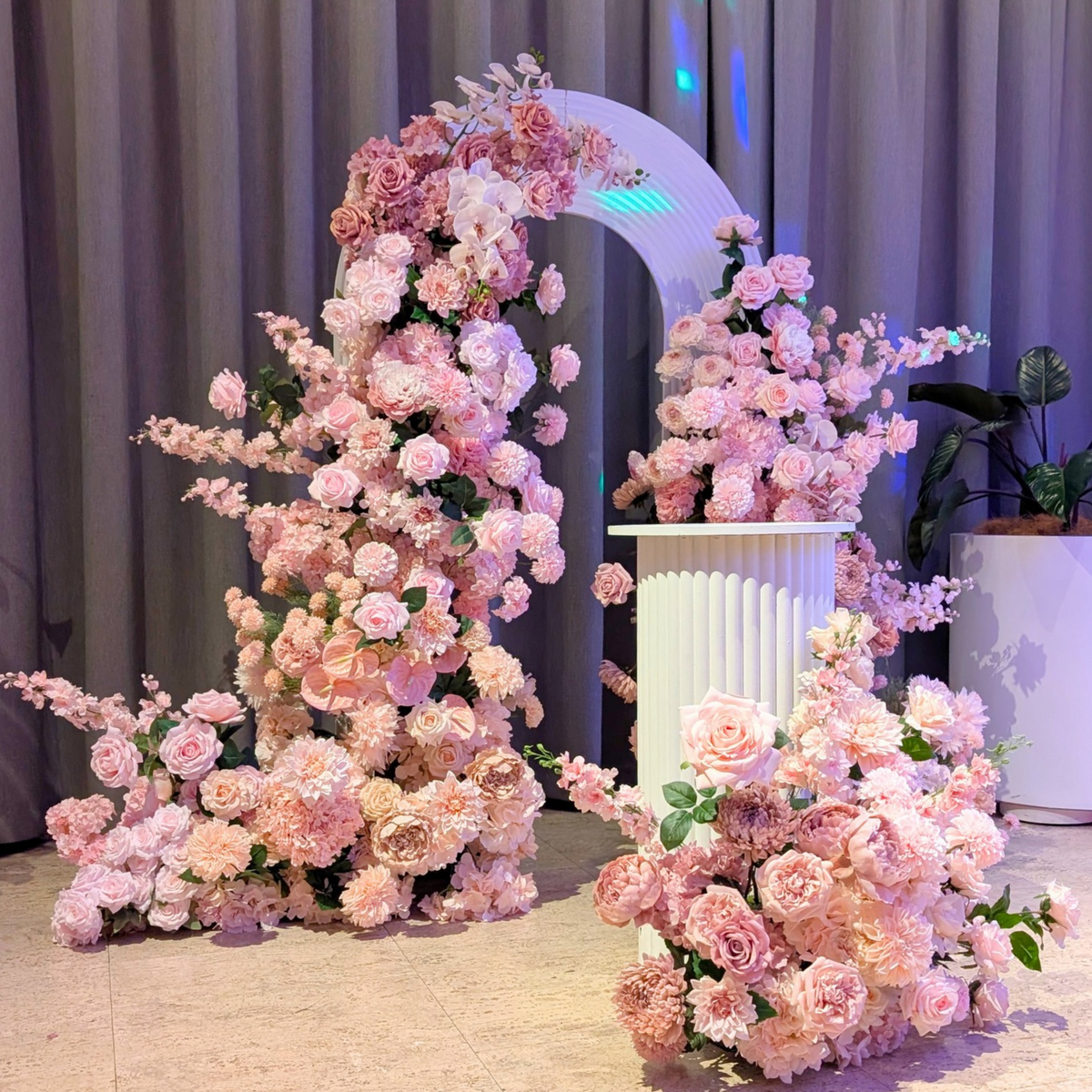 A decorative floral arrangement set featuring large clusters of pink and blush roses with green foliage, styled around an arched backdrop and a white pedestal in an indoor event space. Styled at the Calile Hotel Fortitude Valley by Lush and Lavish Events a Birsbane and Gold Coast based event planning, event styling and on-the-day coordination business.