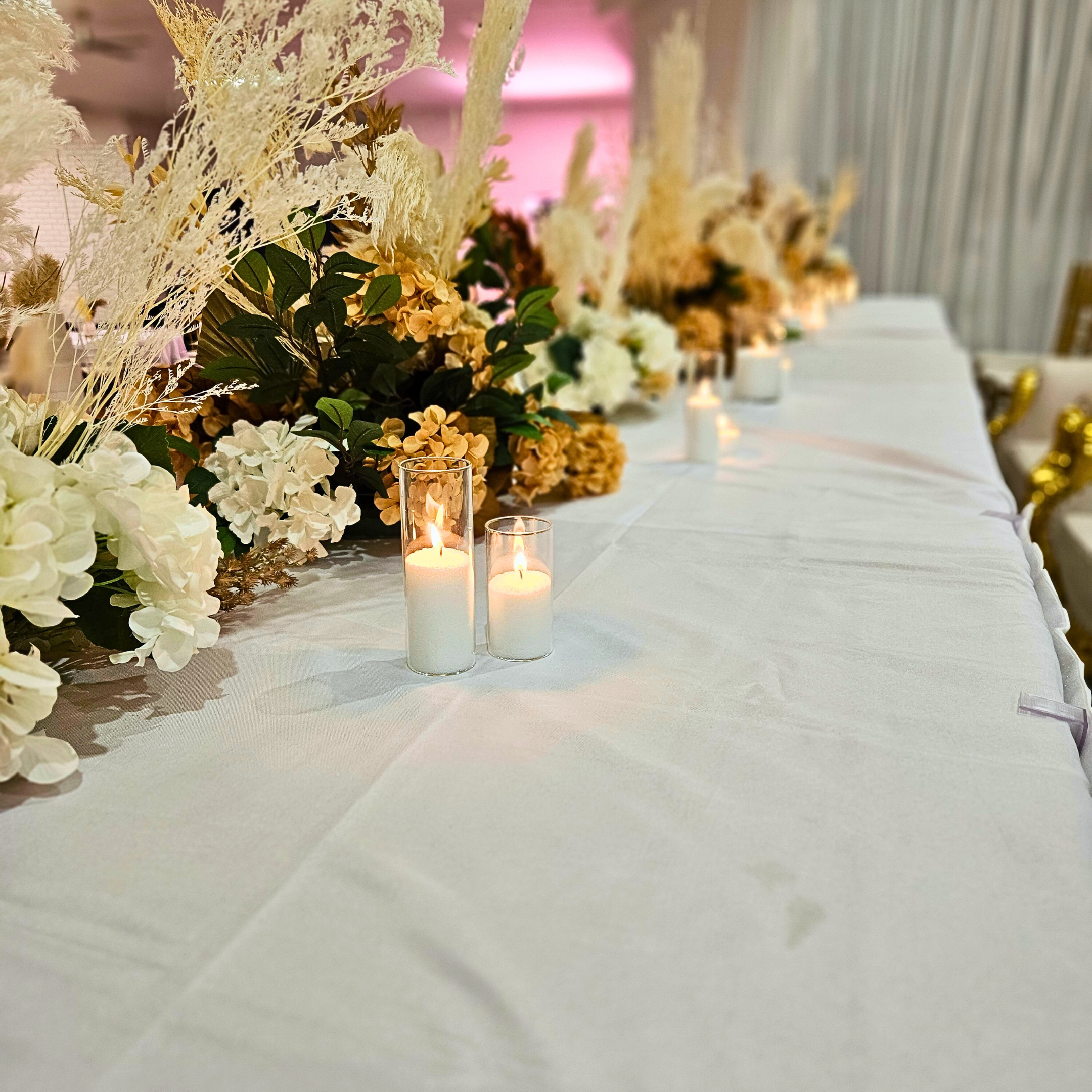 Bridal table with white and gold flowers, tall pampas grass, lit candles, and a white tablecloth. Indoor venue with soft pink lighting. Prop hire, Prop hiring, Prop hire, event stylists Brisbane, brisbane event styling, brisbane event styling, event stylist Brisbane, Brisbane Wedding, Gold Coast Wedding, Floral Styling, Lush & Lavish Events, Brisbane Tablescape, Gold Coast Tablescapes, Brisbane Candle Hire