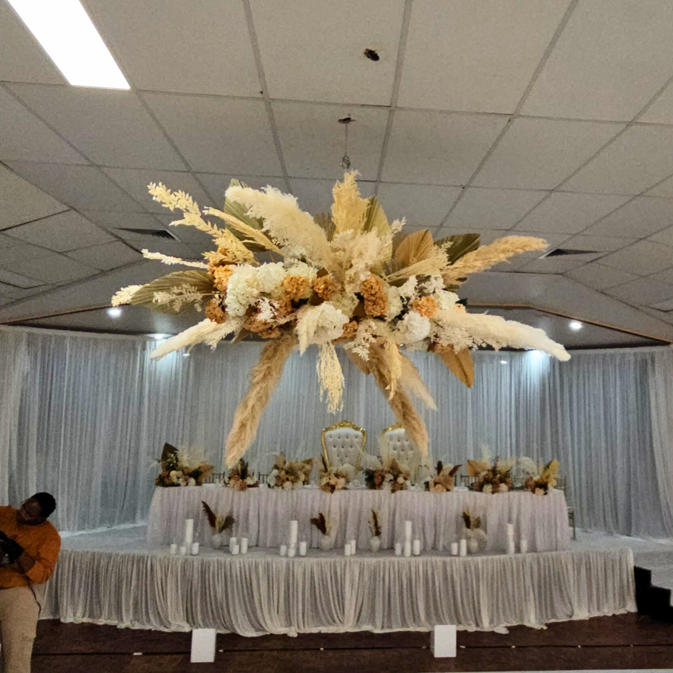 Ornate dried floral arrangement with white and pale yellow flowers, pampas grass, and foliage, suspended above a decorated table against a white backdrop in an event hall. Prop hire, Prop hiring, Prop hire, event stylists Brisbane, brisbane event styling, brisbane event styling, event stylist Brisbane, Brisbane Wedding, Gold Coast Wedding, Floral Styling, Lush & Lavish Events, Brisbane Tablescape, Gold Coast Tablescapes, Brisbane Candle Hire