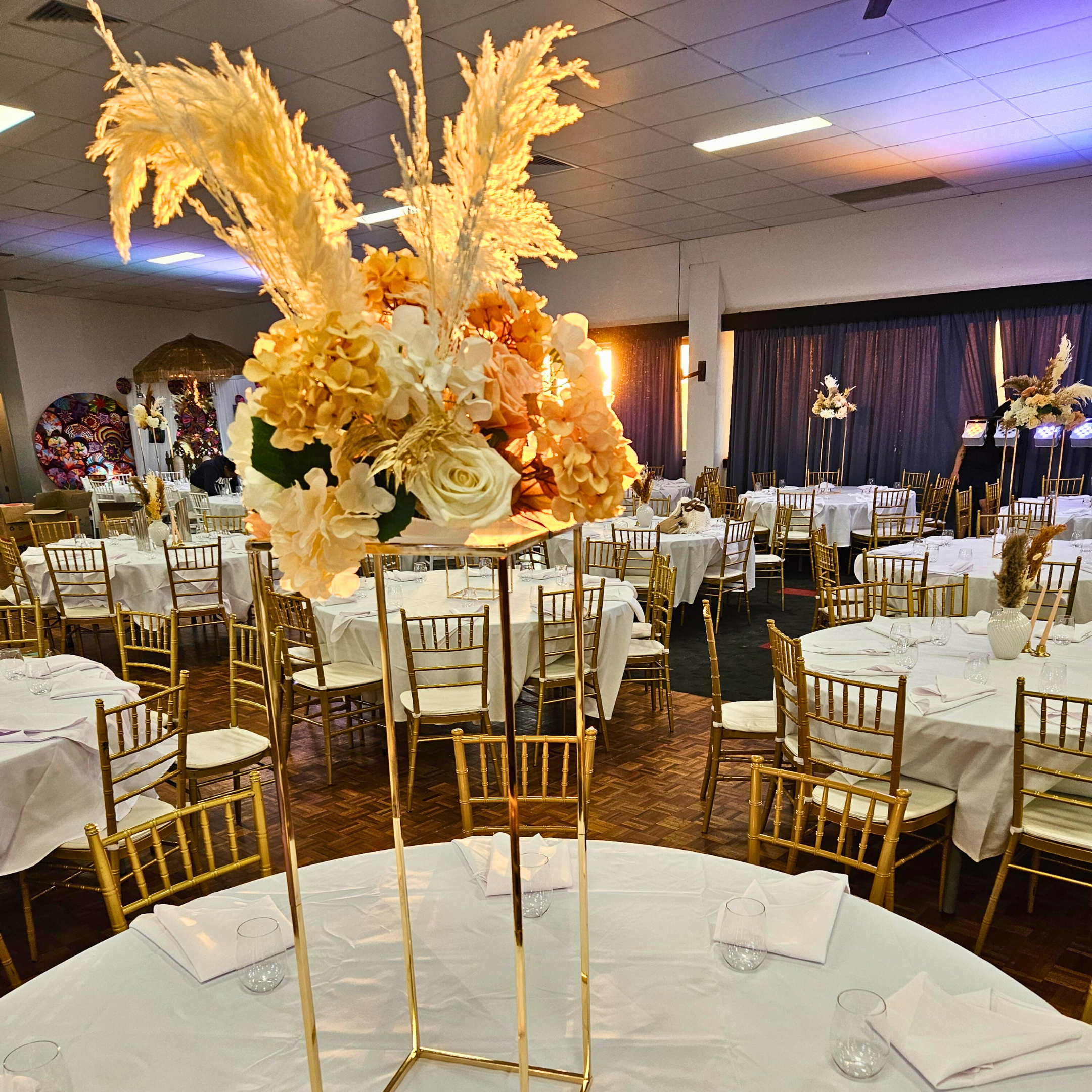 Elegant floral arrangement with white and peach flowers and tall plumes atop a gold plinth, serving as a centerpiece in an event space with round tables set with white linens. Prop hire, Prop hiring, Prop hire, event stylists Brisbane, brisbane event styling, brisbane event styling, event stylist Brisbane, Brisbane Wedding, Gold Coast Wedding, Floral Styling, Lush & Lavish Events, Brisbane Tablescape, Gold Coast Tablescapes, Brisbane Candle Hire