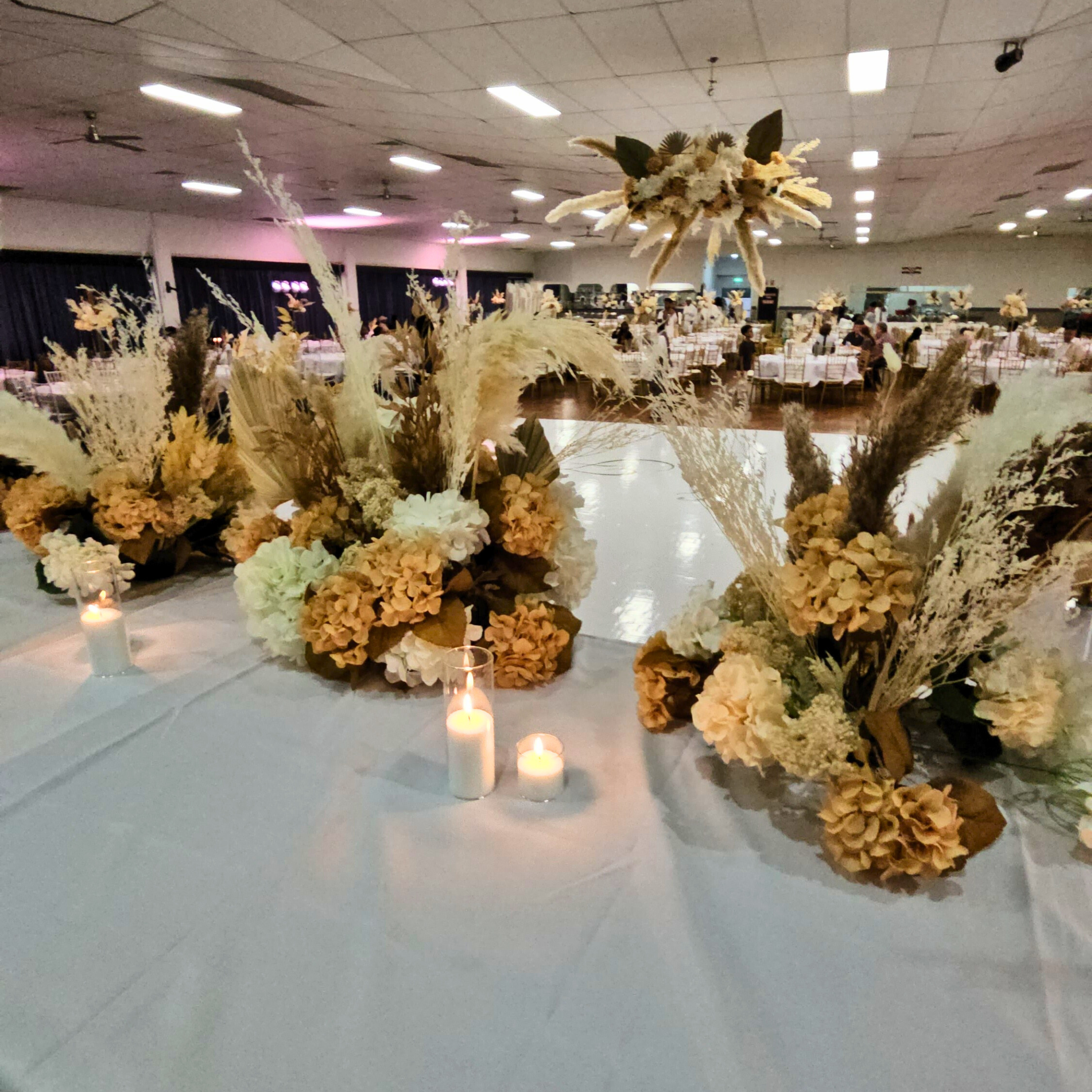 View from the bridal table with cream and pale yellow floral centerpieces, tall pampas grass, and lit candles, creating a warm ambiance. Background shows guest tables with similar decorations and soft lighting. Prop hire, Prop hiring, Prop hire, event stylists Brisbane, brisbane event styling, brisbane event styling, event stylist Brisbane, Brisbane Wedding, Gold Coast Wedding, Floral Styling, Lush & Lavish Events, Brisbane Tablescape, Gold Coast Tablescapes, Brisbane Candle Hire