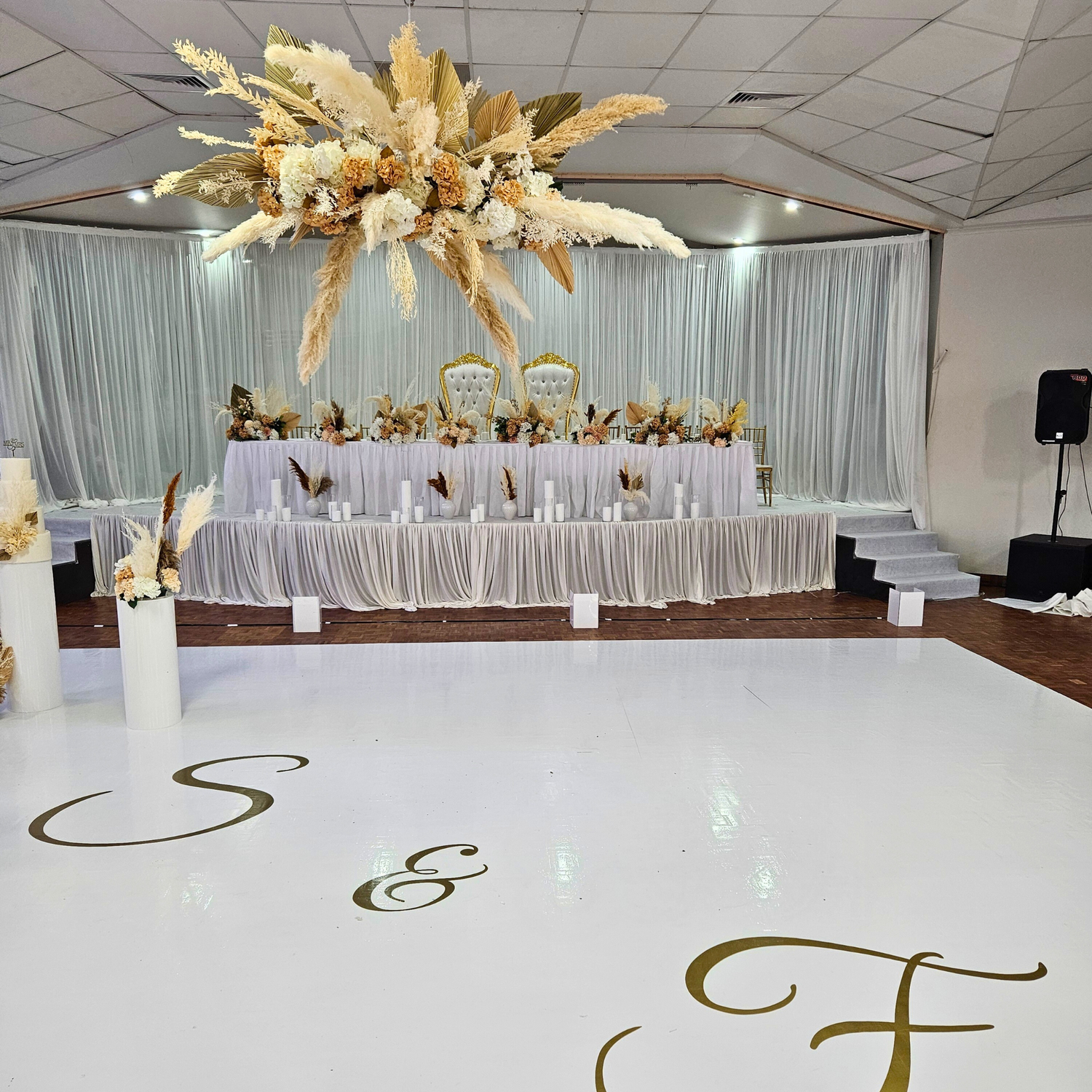 Suspended floral arrangement with white and pale yellow flowers, greenery, and gold accents above a glossy white dance floor with golden initials. Background shows a head table with matching decorations and silvery curtains. Prop hire, Prop hiring, Prop hire, event stylists Brisbane, brisbane event styling, brisbane event styling, event stylist Brisbane, Brisbane Wedding, Gold Coast Wedding, Floral Styling, Lush & Lavish Events, Brisbane Tablescape, Gold Coast Tablescapes, Brisbane Candle Hire