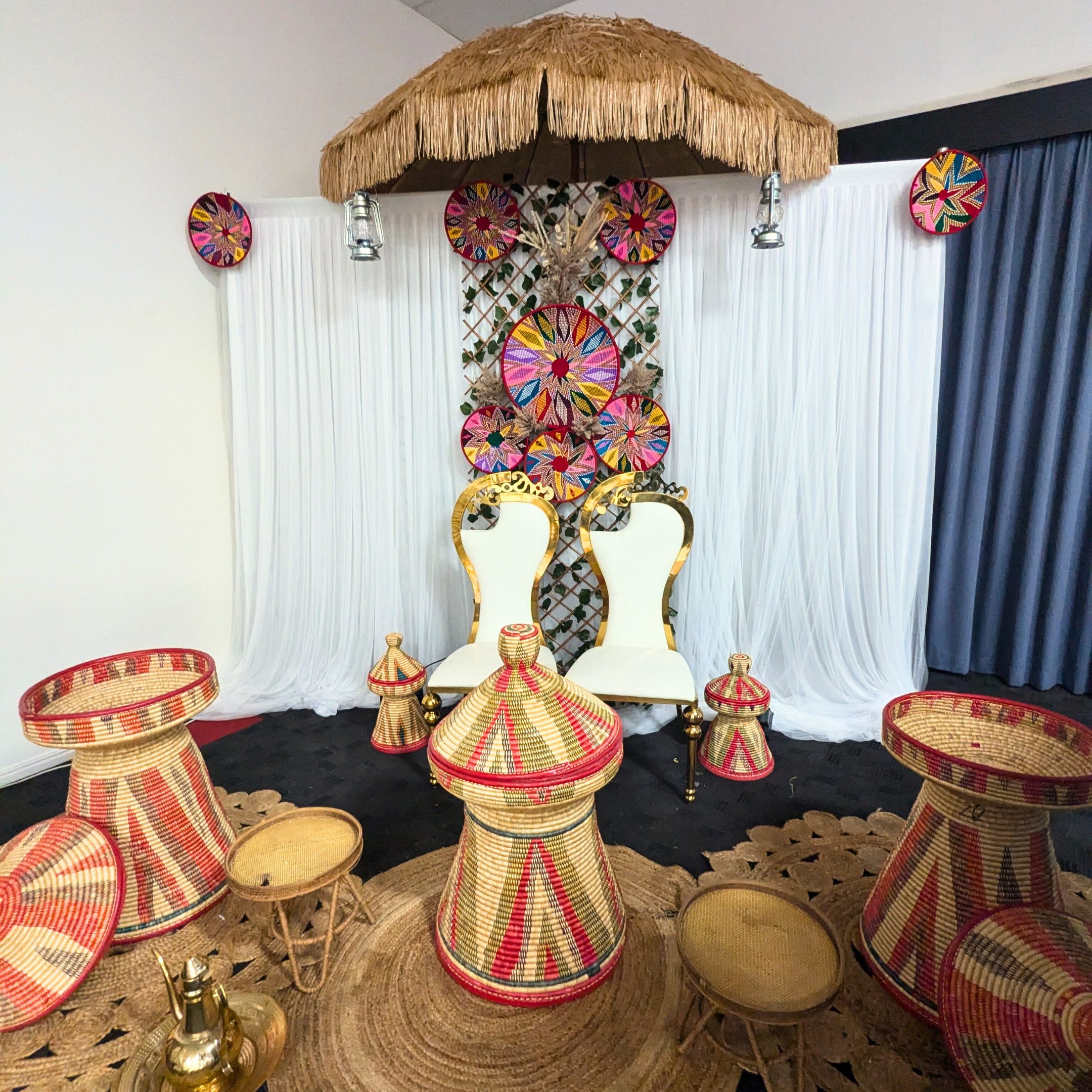 Traditional Ethiopian setup with a double chair with high woven backrest, white curtains, colorful circular patterns on the wall, thatched roof decoration, handwoven baskets and stools, and a small brass coffee pot. Prop hire, Prop hiring, Prop hire, event stylists Brisbane, brisbane event styling, brisbane event styling, event stylist Brisbane, Brisbane Wedding, Gold Coast Wedding, Floral Styling, Lush & Lavish Events, Brisbane Tablescape, Gold Coast Tablescapes, Brisbane Candle Hire